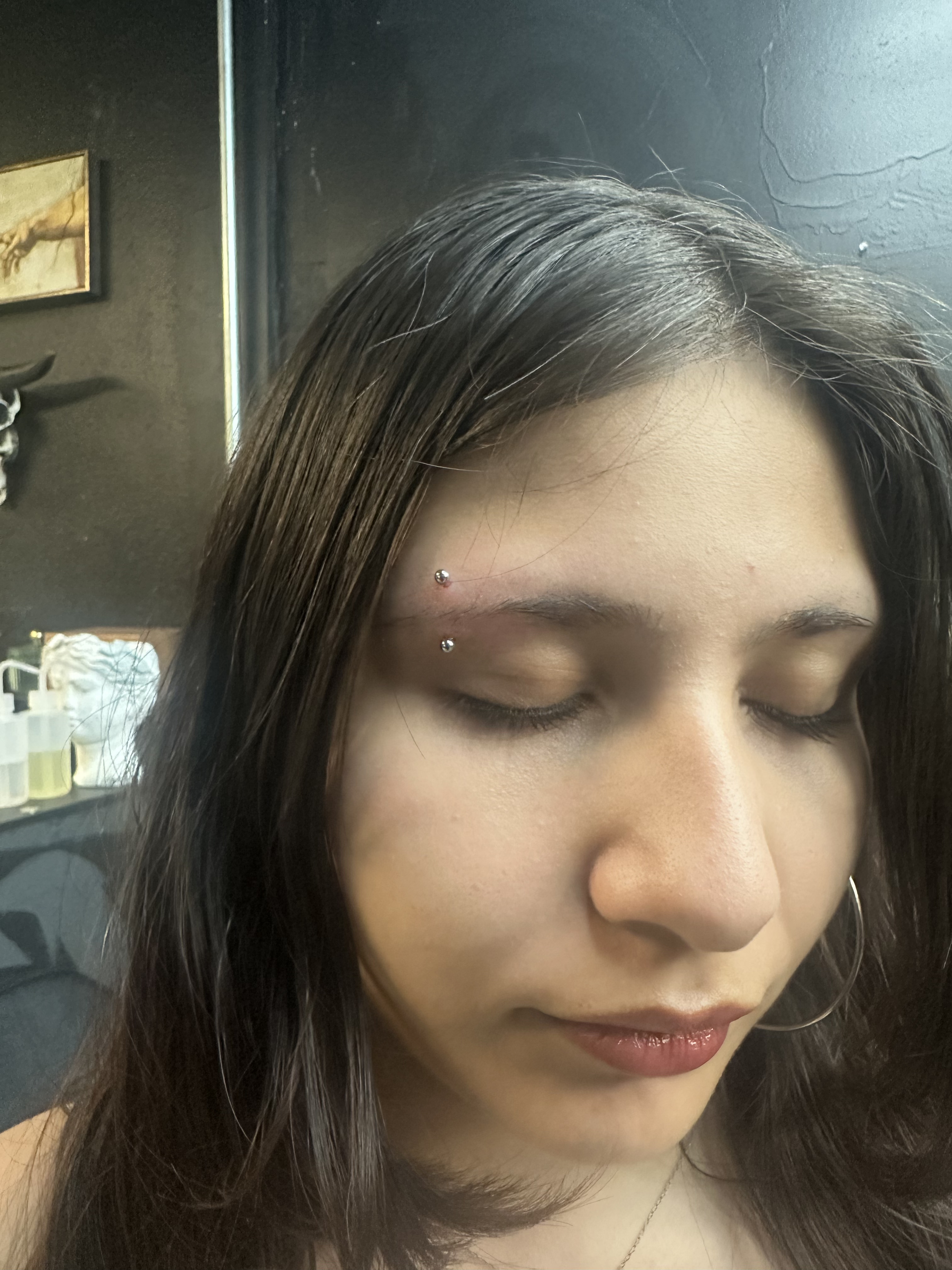 A young woman with dark brown hair and hoop earrings, with her eyes closed, in a room with black walls and a framed picture of the hand of God.