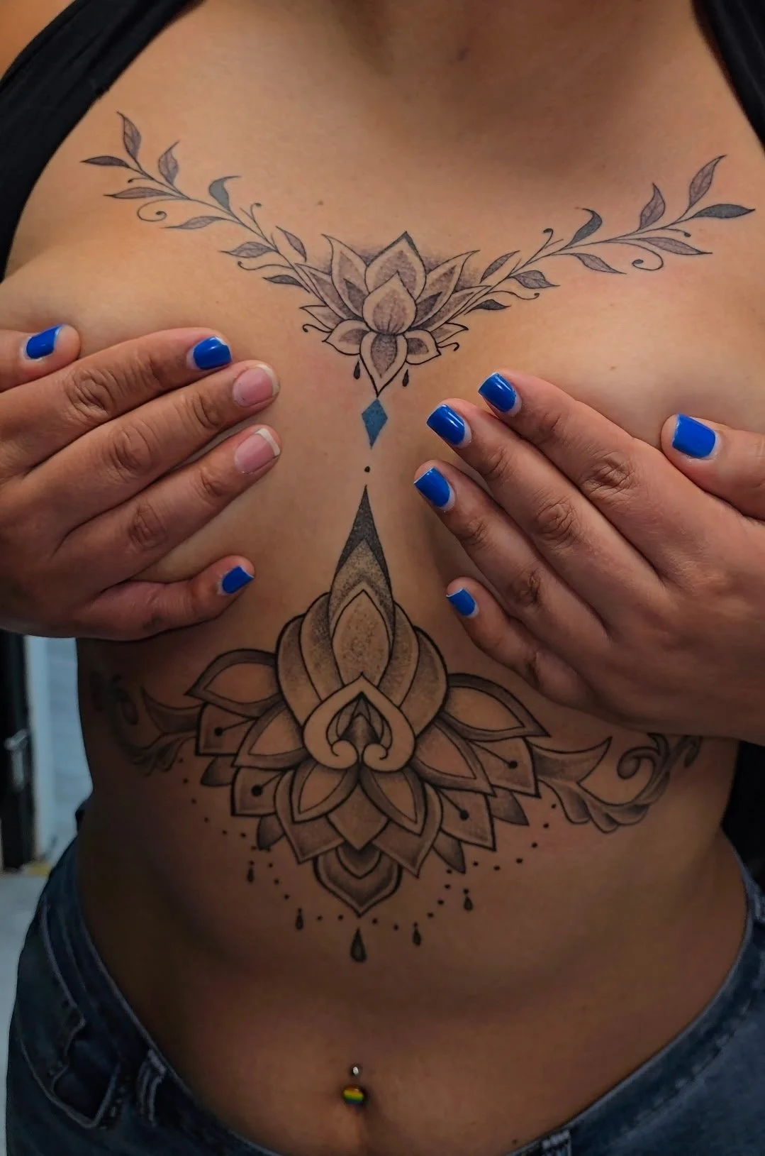 Close-up of a tattoo on a person's chest and stomach, featuring a symmetrical floral and geometric design. The person has hands with blue nail polish covering part of the tattoo, and has a rainbow-colored belly button piercing.