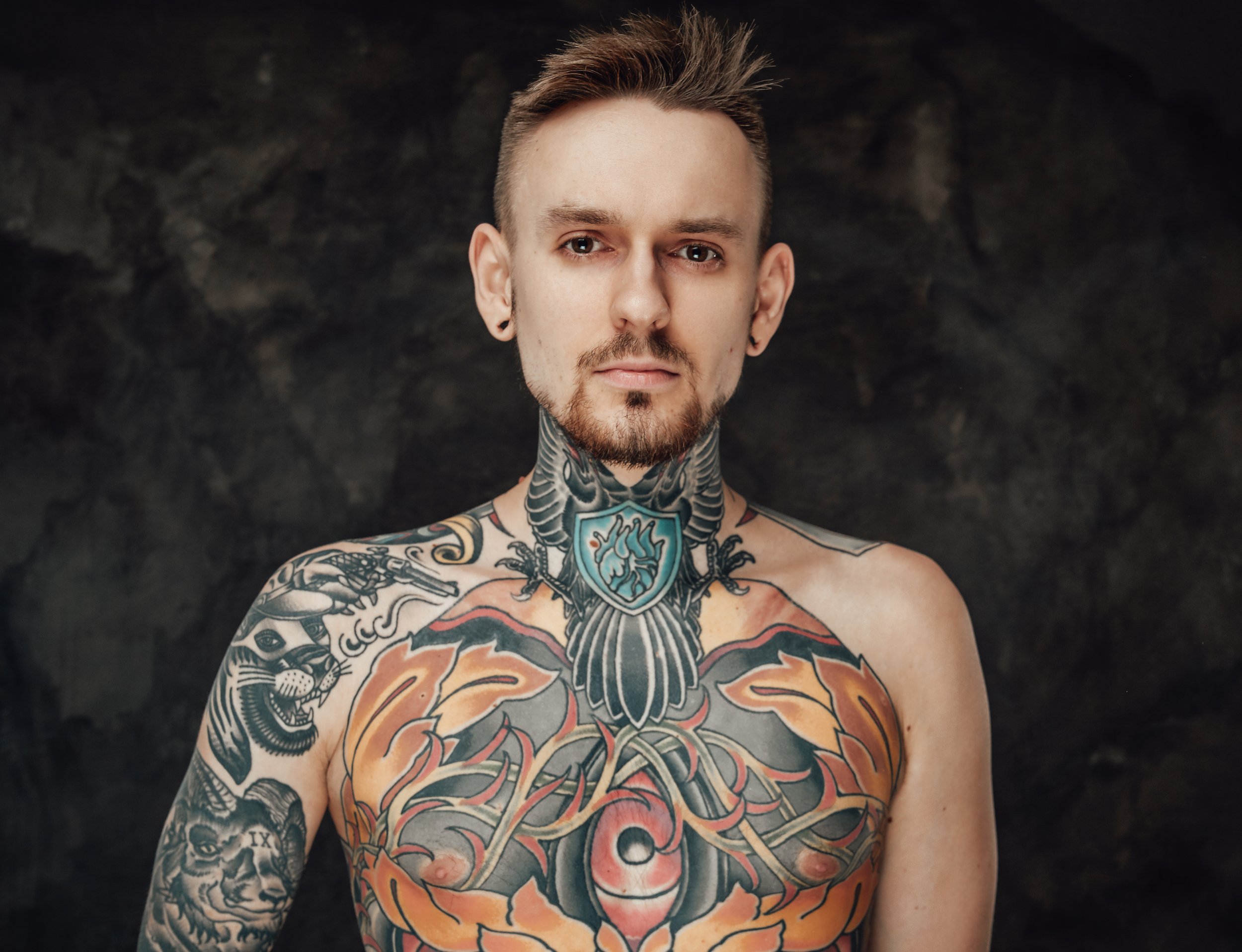 A shirtless man with extensive tattoos on his chest and arms, and a tattoo on his neck, standing against a dark background.