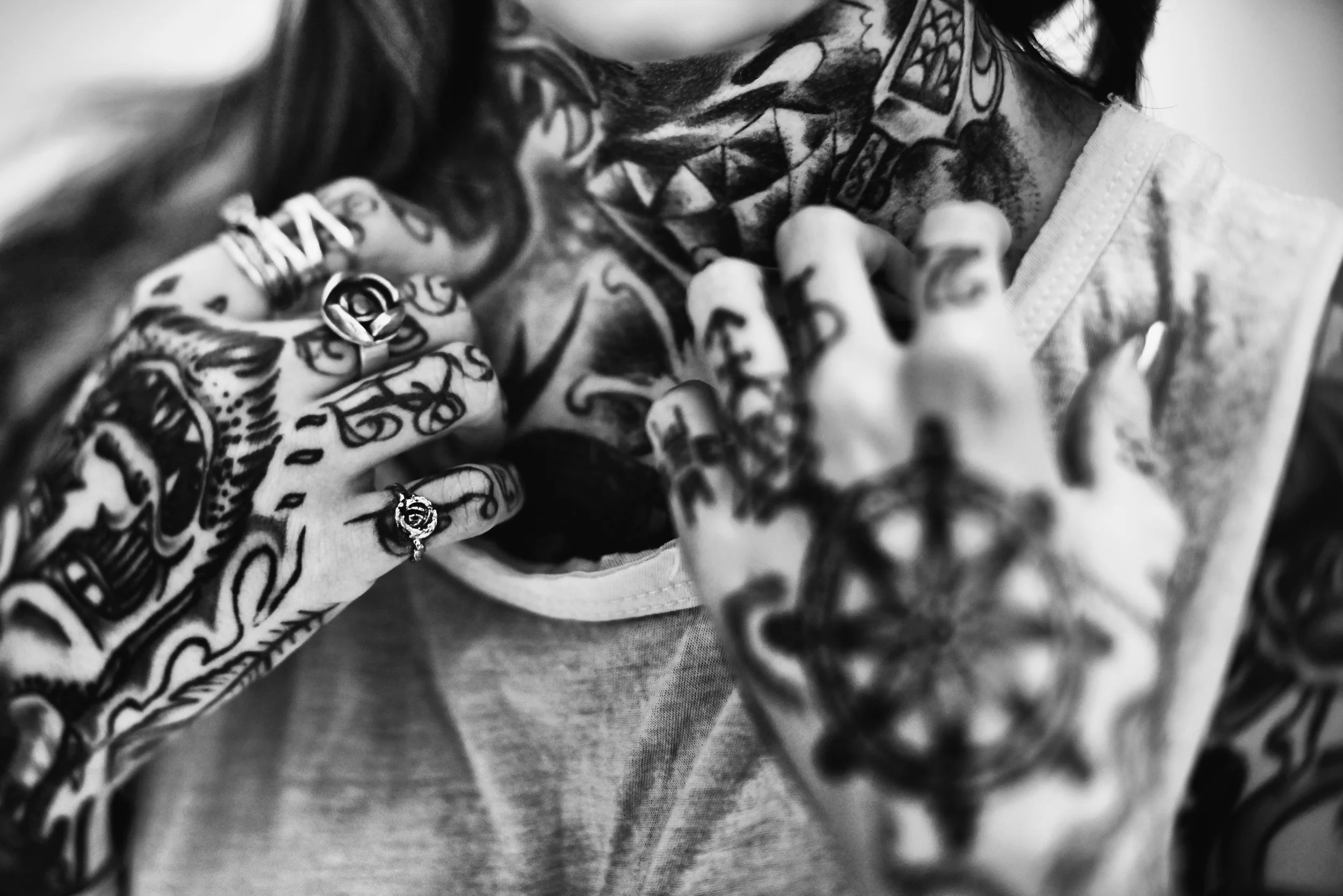 Black and white photo of a person with multiple tattoos on their arms and hands, wearing rings, holding and kissing a skull that also has tattoos.
