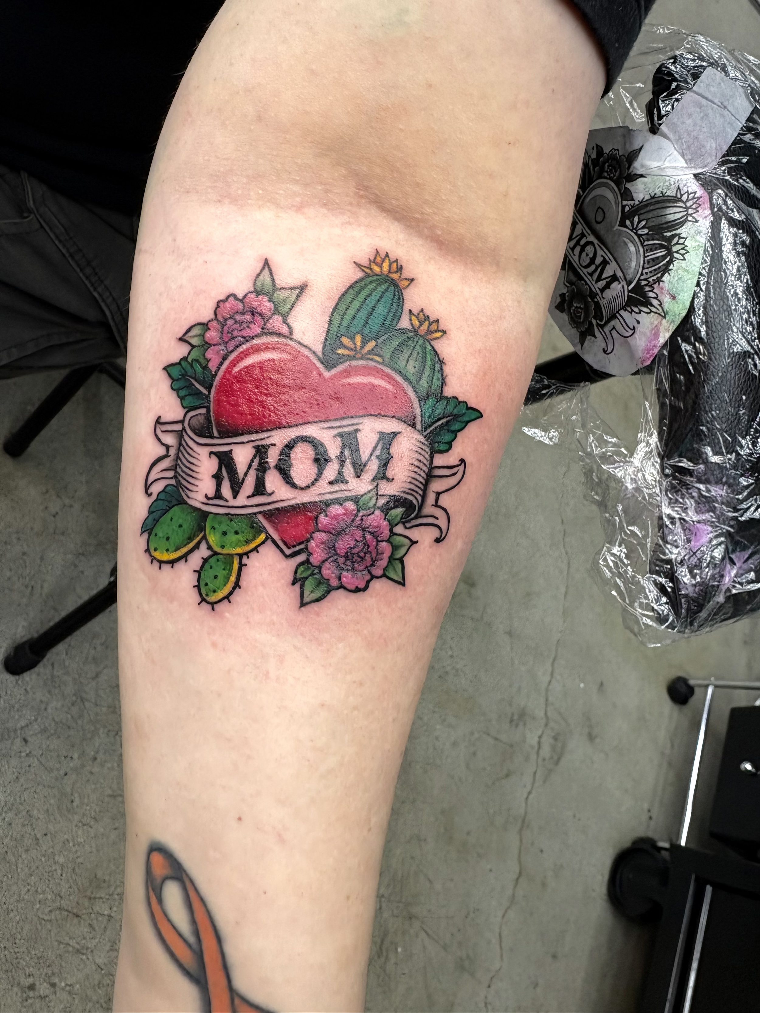 A colorful tattoo on a person's thigh featuring a heart with the word 'MOM' written across a banner, surrounded by cacti and pink flowers.