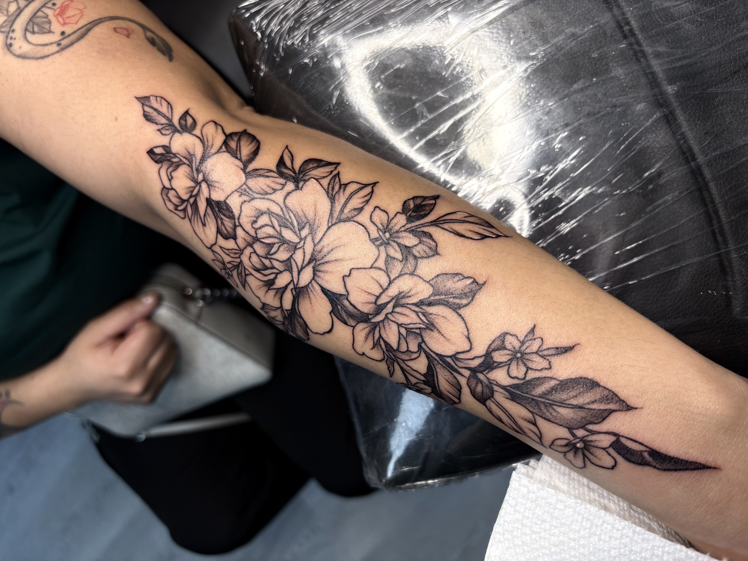 A person's forearm with a black outline floral tattoo featuring large flowers and leaves, resting on a plastic-covered surface.