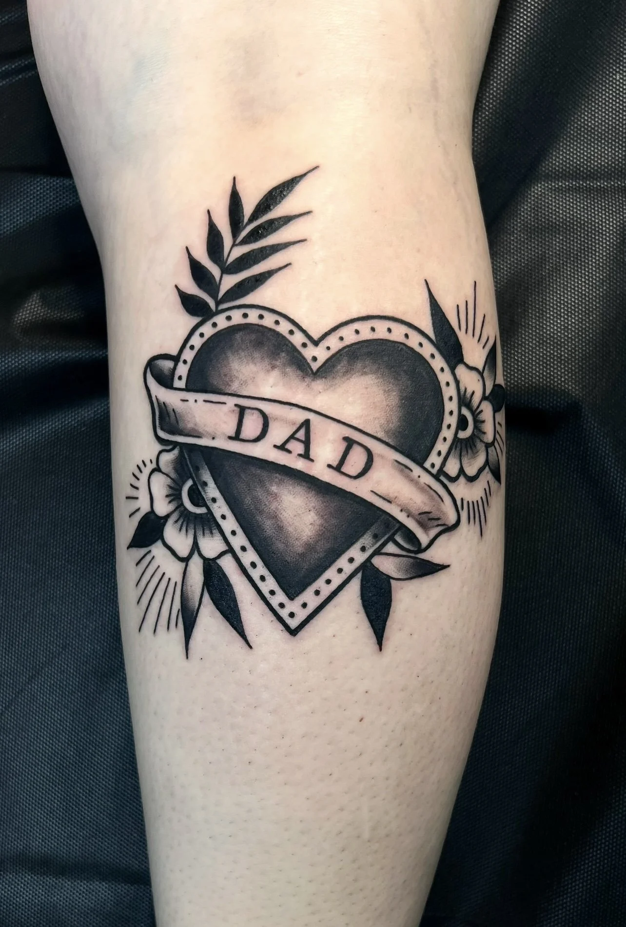 Tattoo of a heart with a banner reading 'DAD', surrounded by flowers, leaves, and decorative lines.