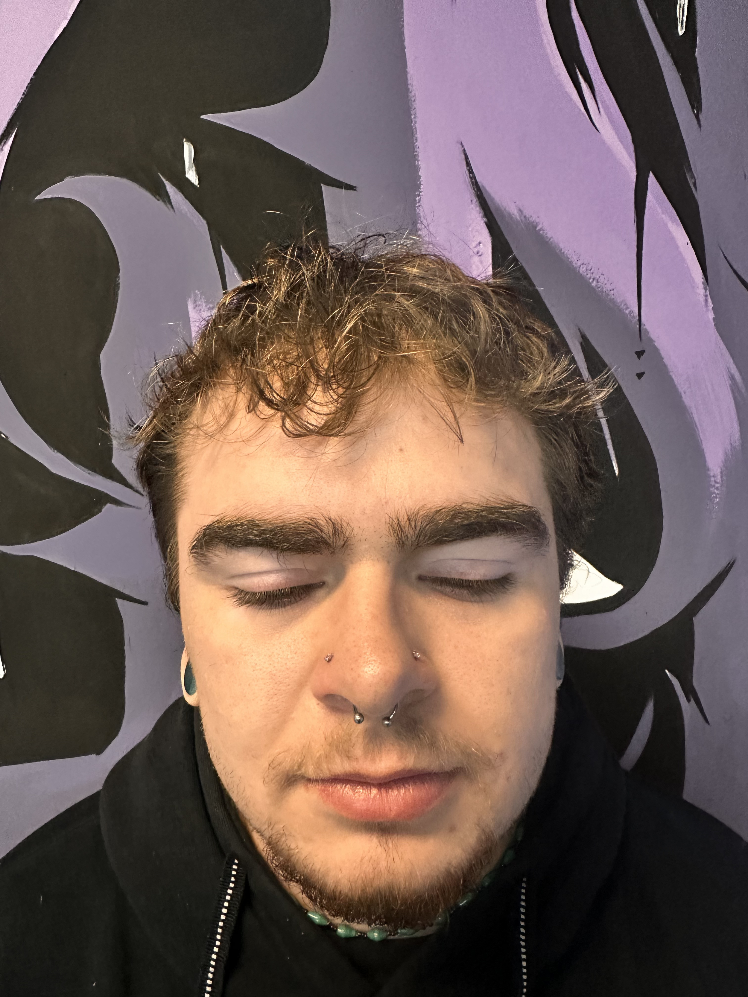 Portrait of a young man with closed eyes, curly hair, multiple facial piercings, and a black hoodie, standing against a purple and black abstract mural background.