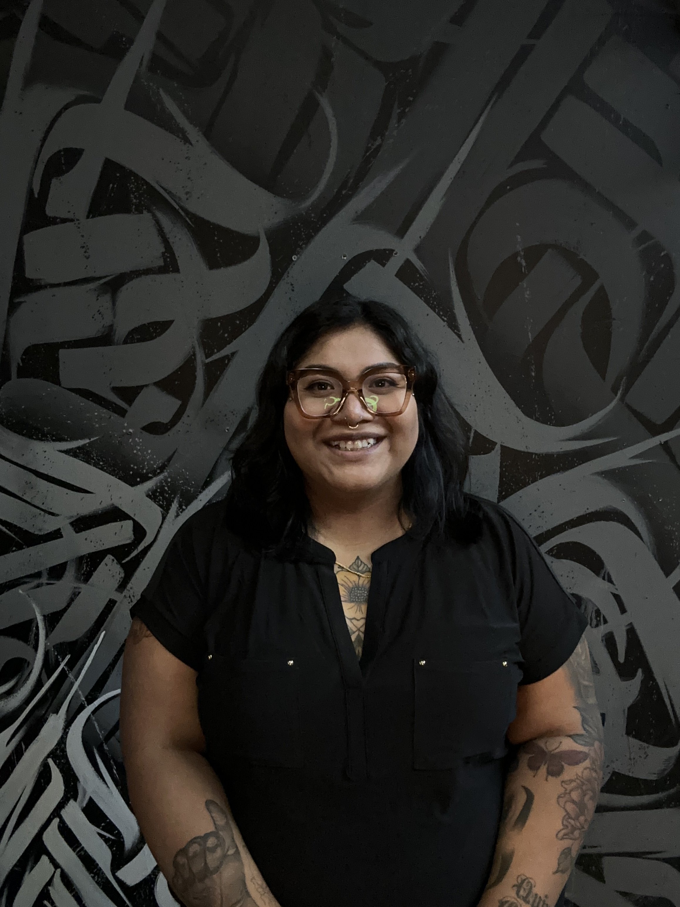 A woman with black hair, glasses, and tattoos on her arms inside a room with a dark, abstract mural on the wall behind her.