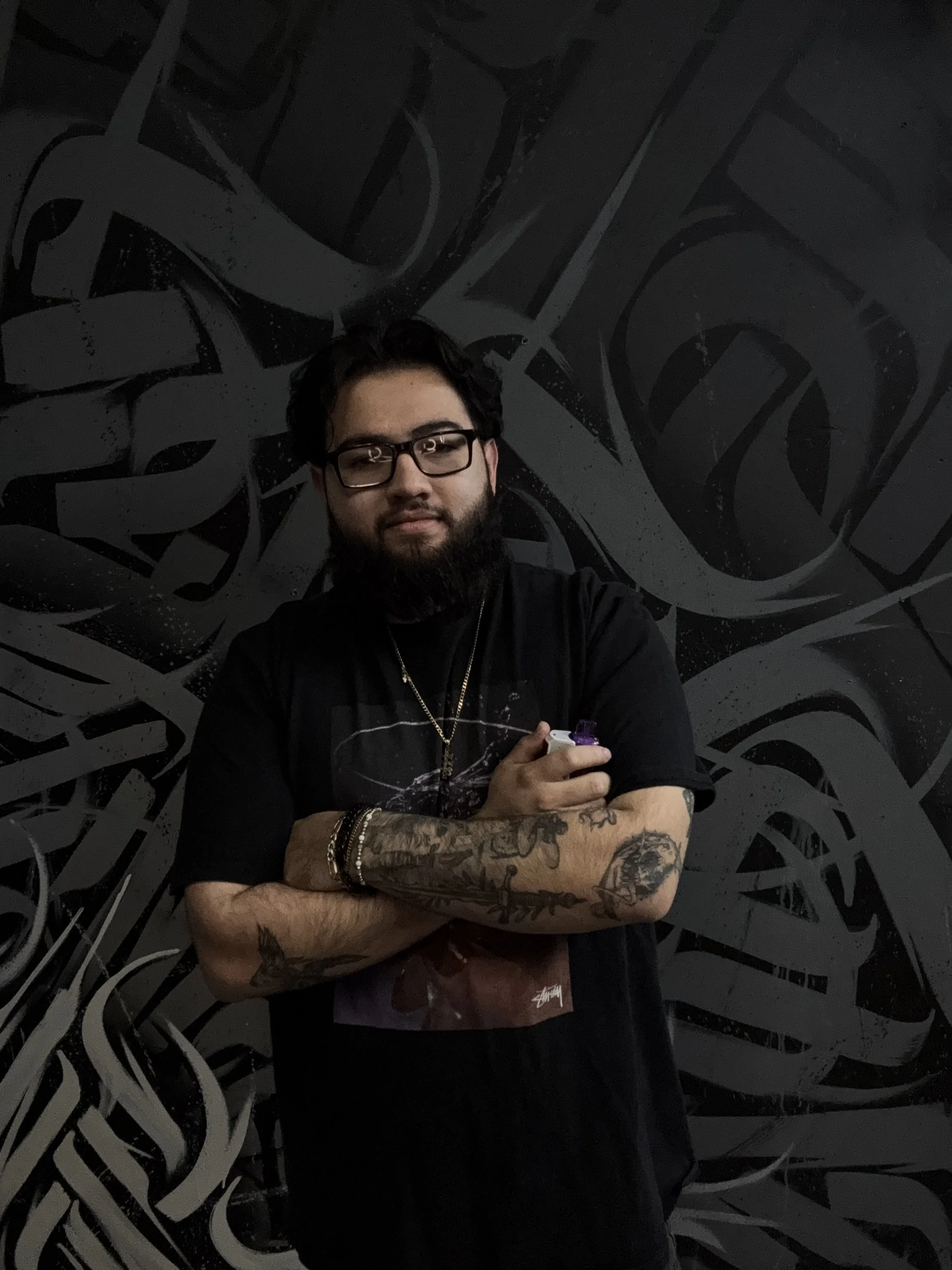 A man with glasses and tattoos on his arms is standing in front of a black wall with graffiti-style art. He has a beard and is holding an object in his right hand.