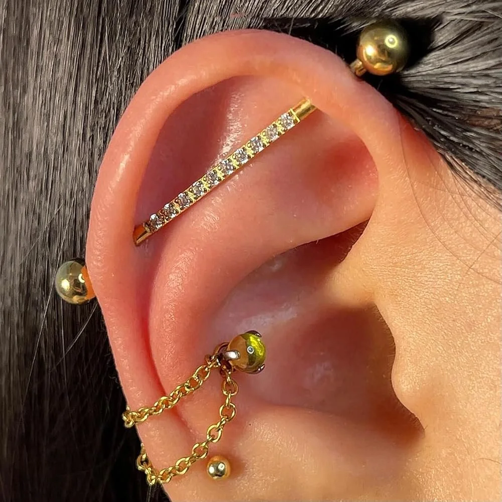Close-up of a person's ear with multiple gold earrings, including a chain connecting two earrings, a hoop with rhinestones, and three plain gold balls.