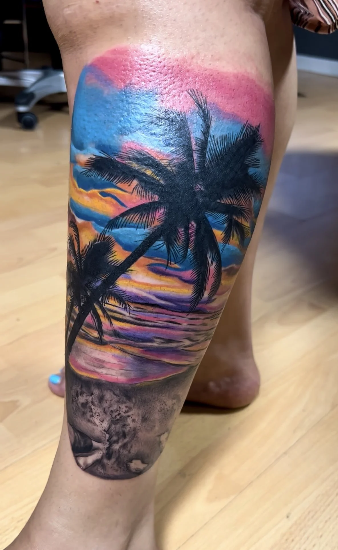 Tattoo of a beach scene with palm trees at sunset or sunrise, featuring colorful sky and water reflections, on a person's calf.
