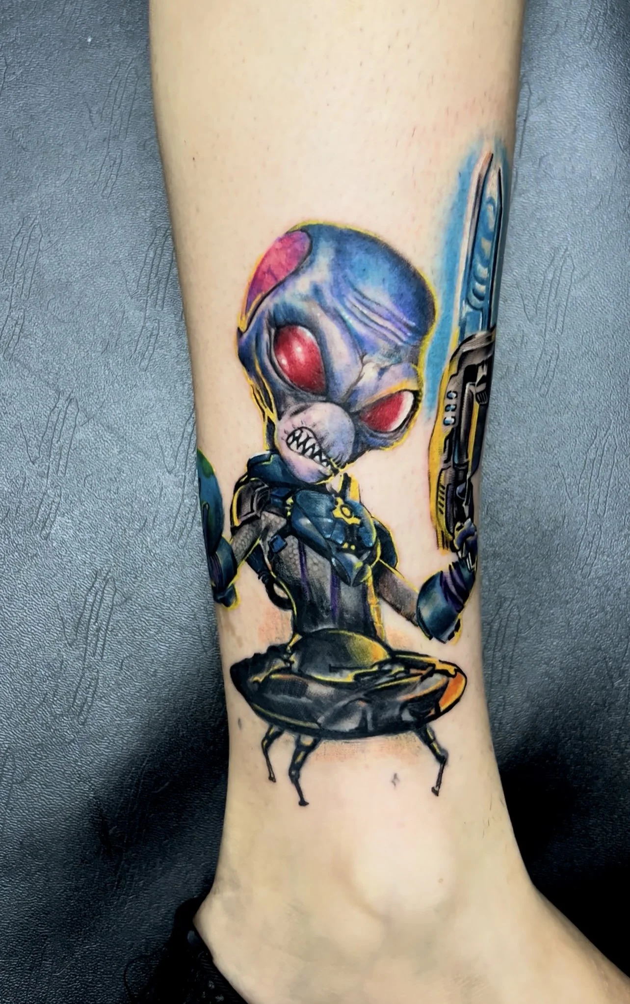 A colorful tattoo of an alien with large red eyes and sharp teeth, holding a large gun, and riding a robotic bug, on a person's forearm.