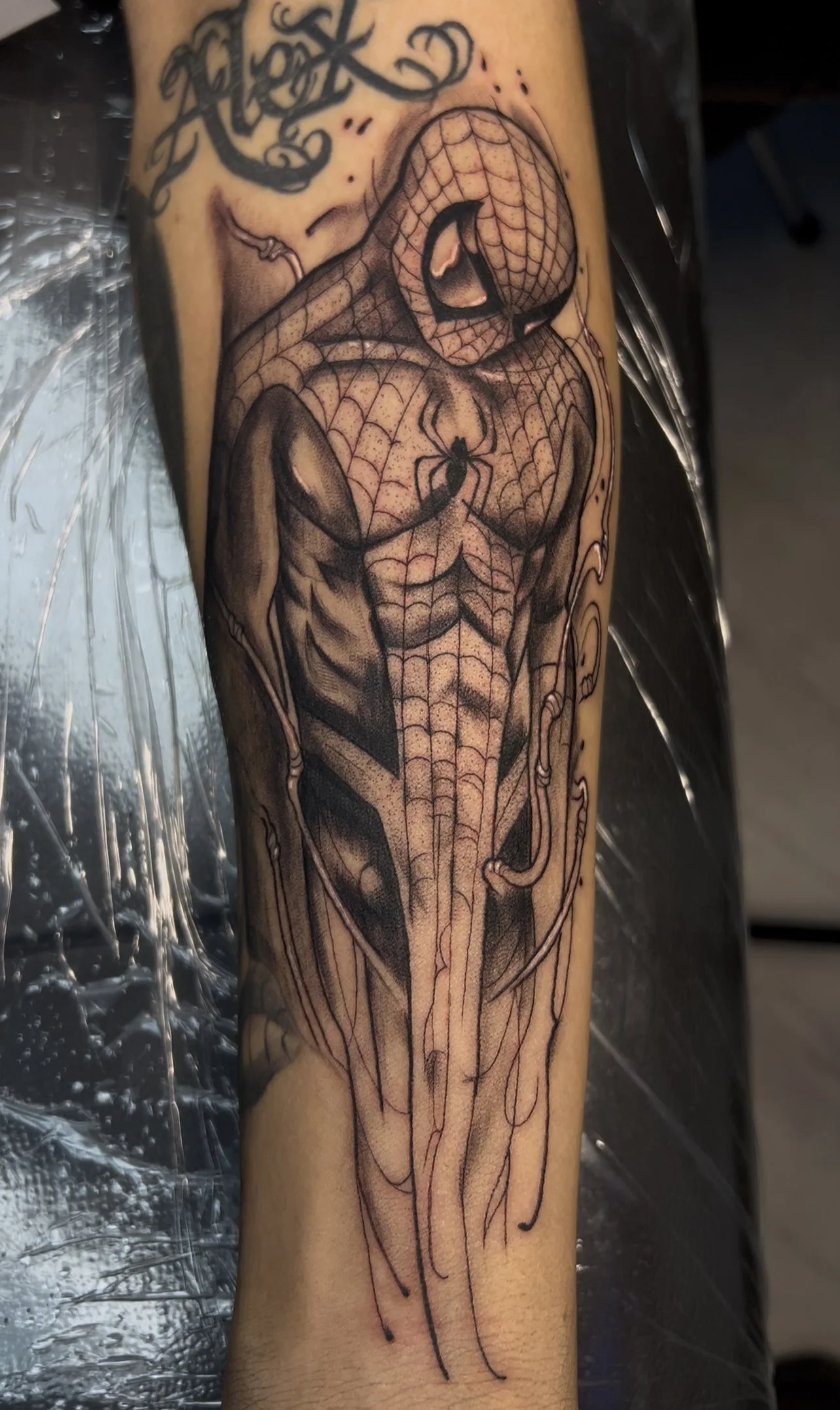 Black and gray tattoo of Spider-Man with vivid web patterns on his suit and a spider emblem on his chest, on upper arm or forearm.