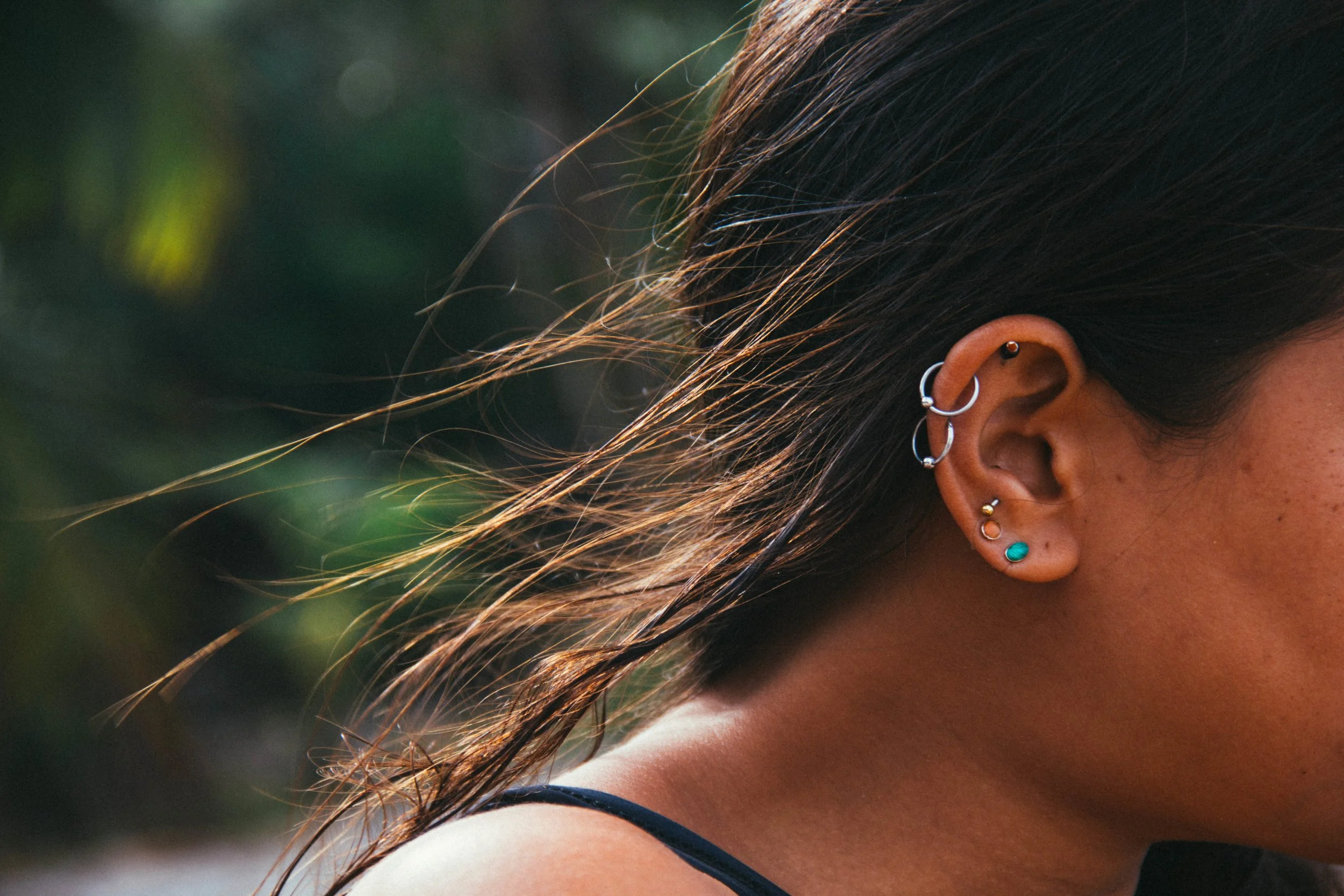 Close-up of a woman's ear with multiple earrings and long brown wavy hair, outdoors with blurred green background.