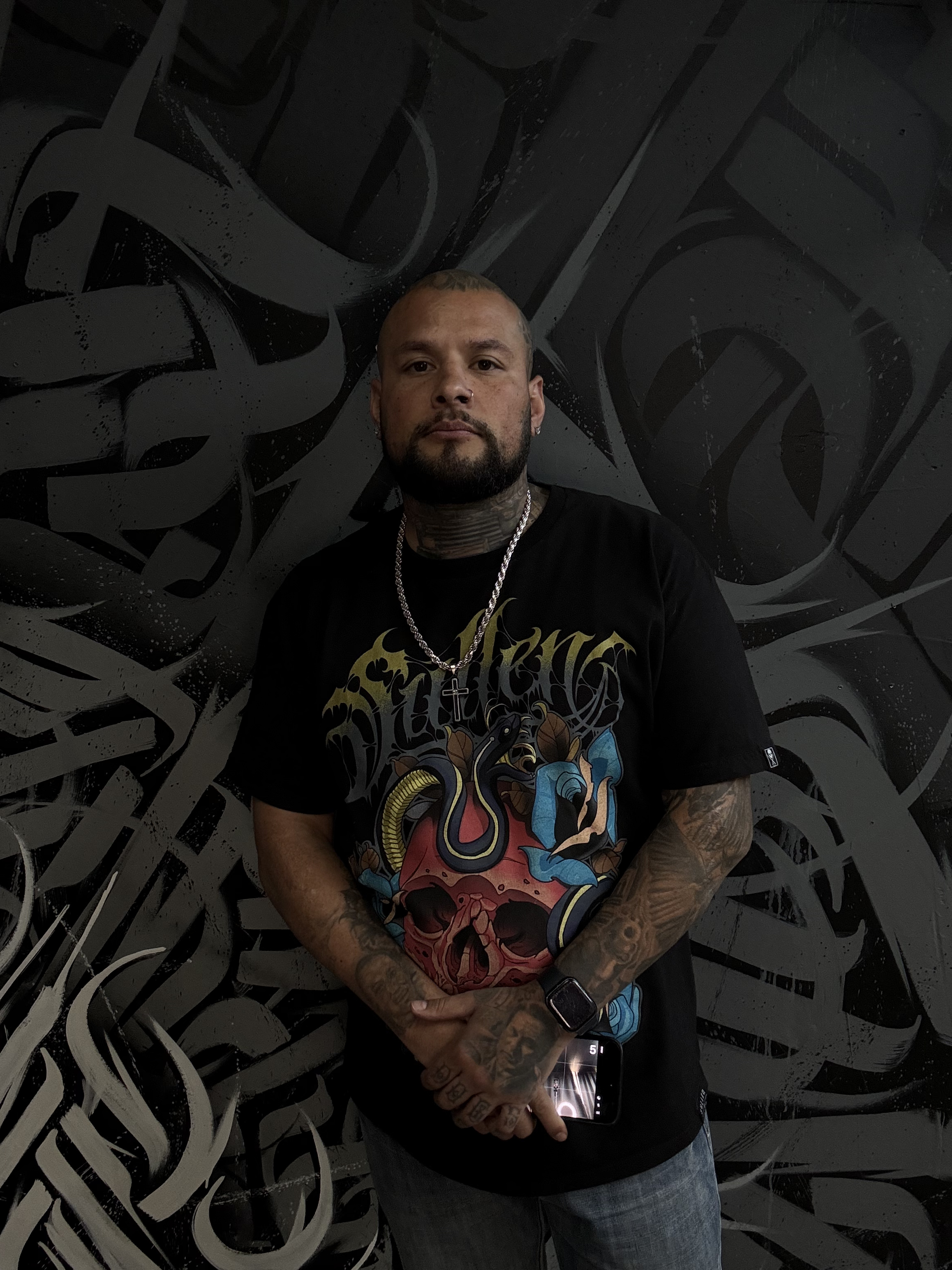 A man with tattoos, a beard, and an earring stands in front of a black and gray graffiti wall. He is wearing a black t-shirt with a colorful graphic design, a silver chain, and a watch. He holds a smartphone in his left hand.