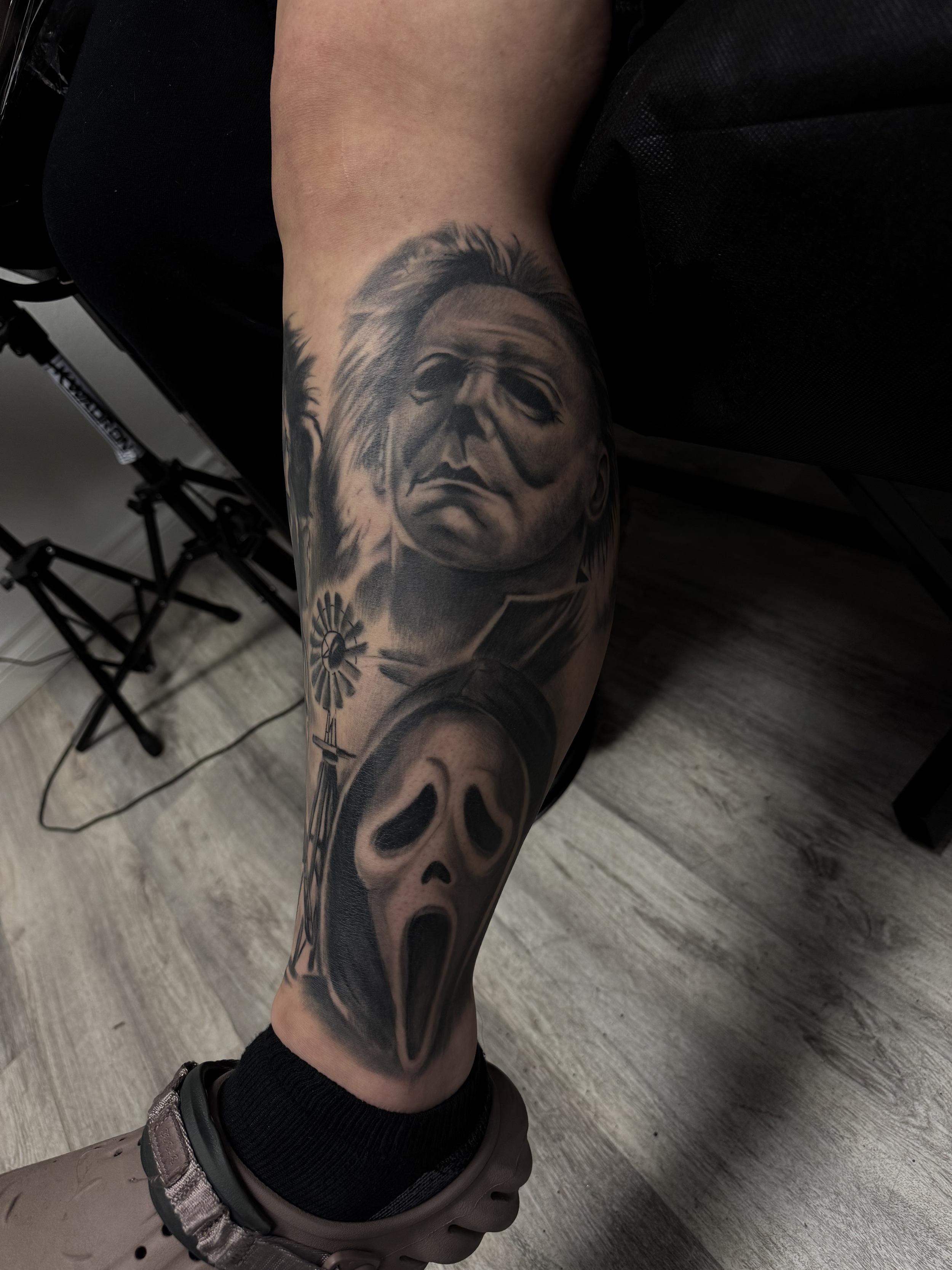 Tattoo of a Micael Meyers, a ghost face mask from the movie Scream, and a windmill on a person's leg.
