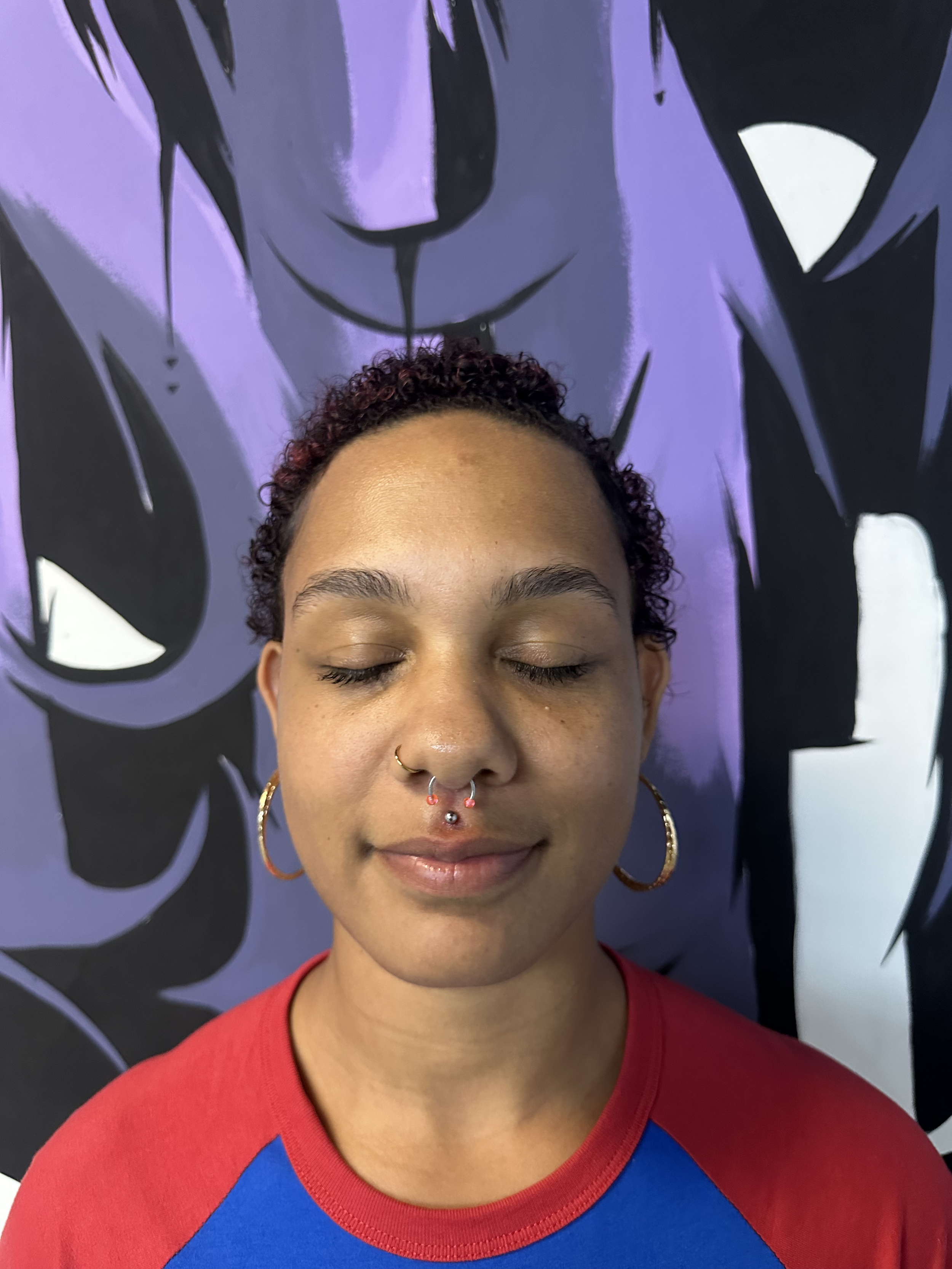A young woman with short curly hair, closed eyes, wearing gold hoop earrings, a septum piercing, and a red and blue shirt, standing in front of a large purple and black abstract mural of a person's face with closed eyes and long black hair.