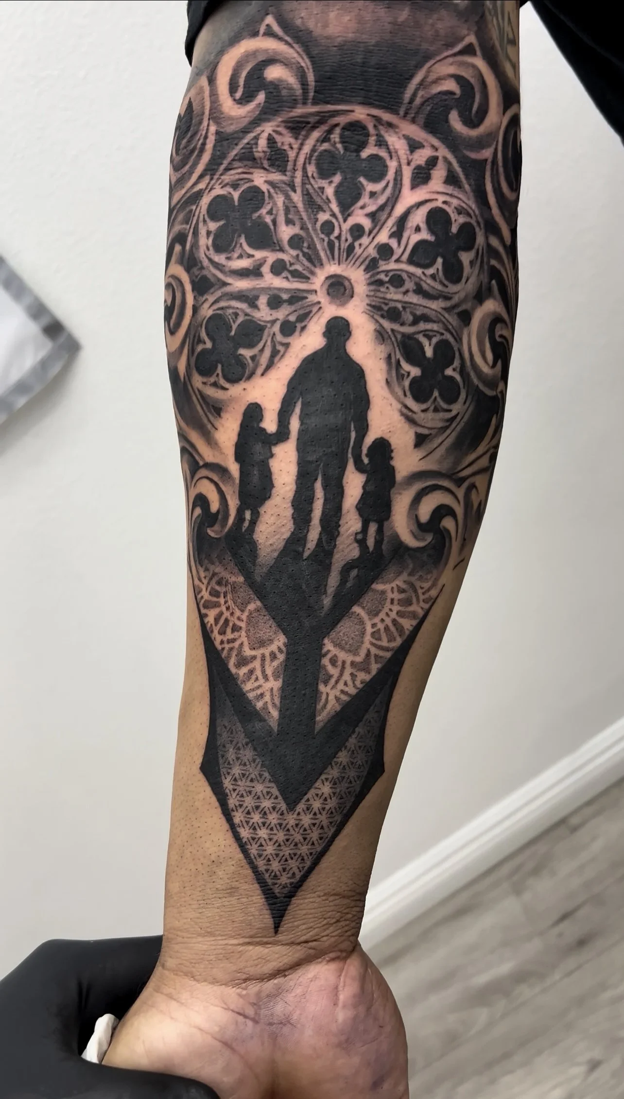 Tattoo of a silhouetted person holding hands with two children, set against a patterned background with floral and geometric designs.