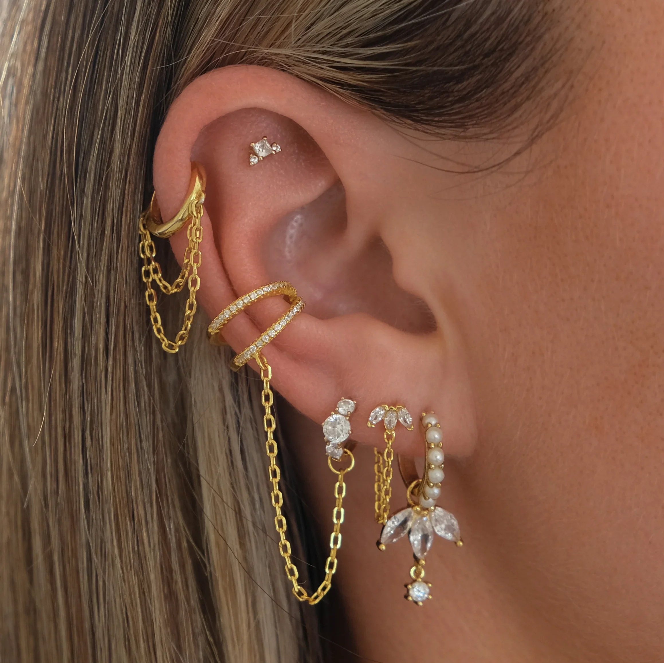 Close-up of a person's ear adorned with multiple gold earrings, including a chain dangle earring, a hoop earring, and a piercing with a small diamond-studded charm.