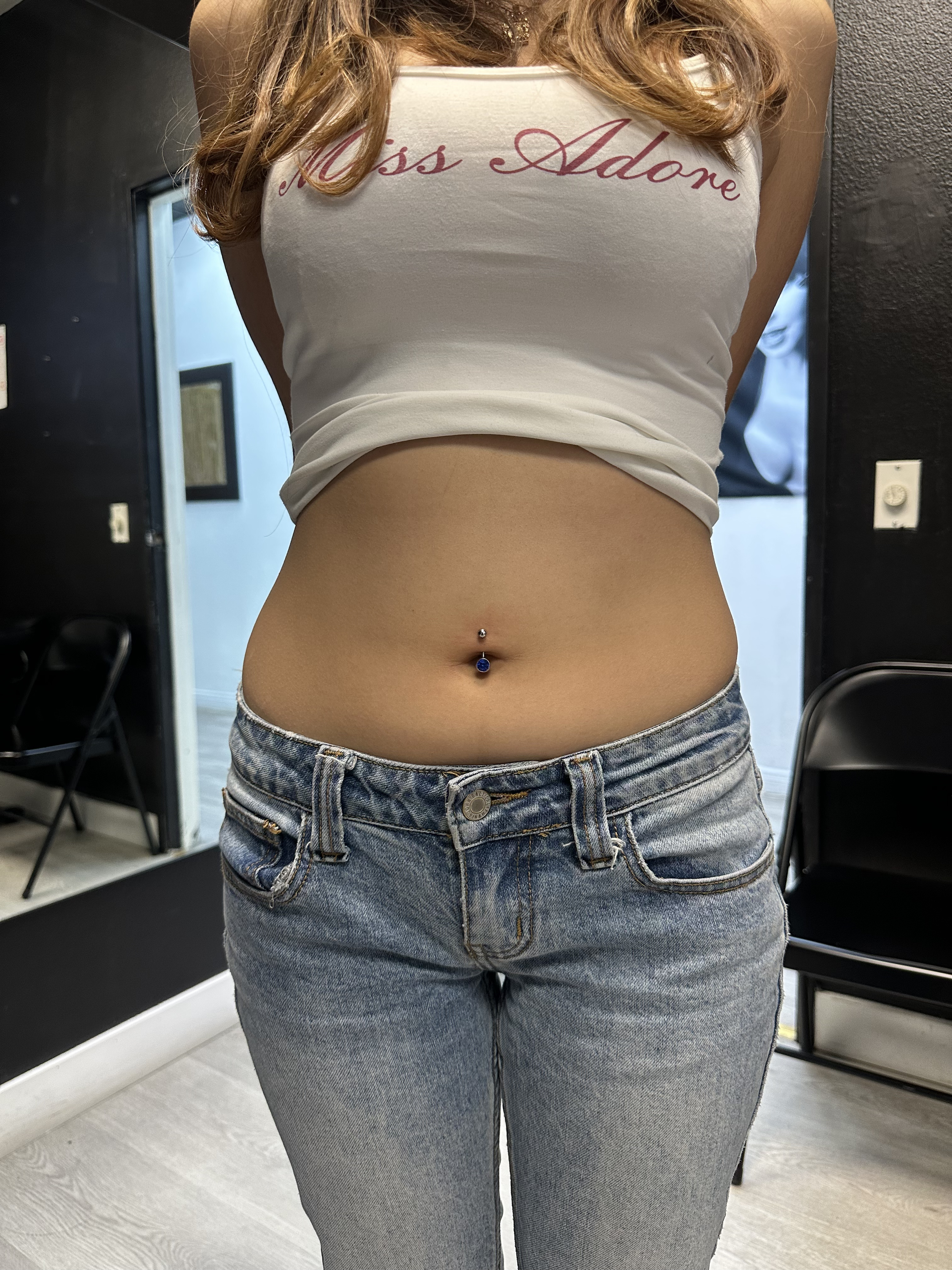 A woman wearing a white tank top with red cursive writing and blue jeans, standing indoors. The top says 'Miss Adone'.