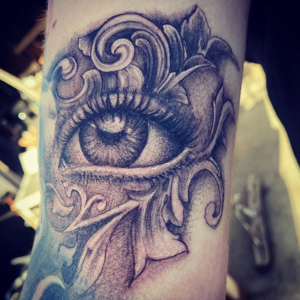 Tattoo of a detailed human eye with decorative swirling patterns around it.