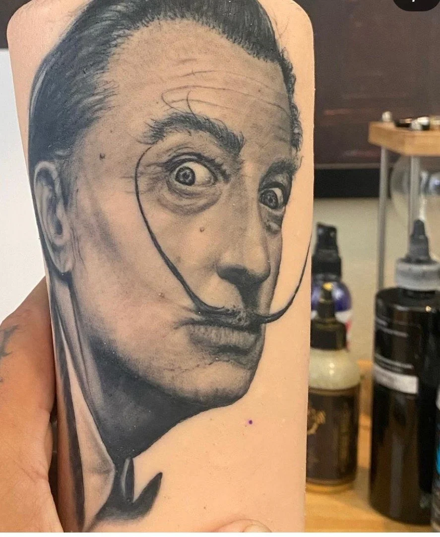 Detailed black and gray tattoo of a vintage portrait of a man with exaggerated facial features, including wide eyes, bushy eyebrows, and a curled mustache, on a person's arm.