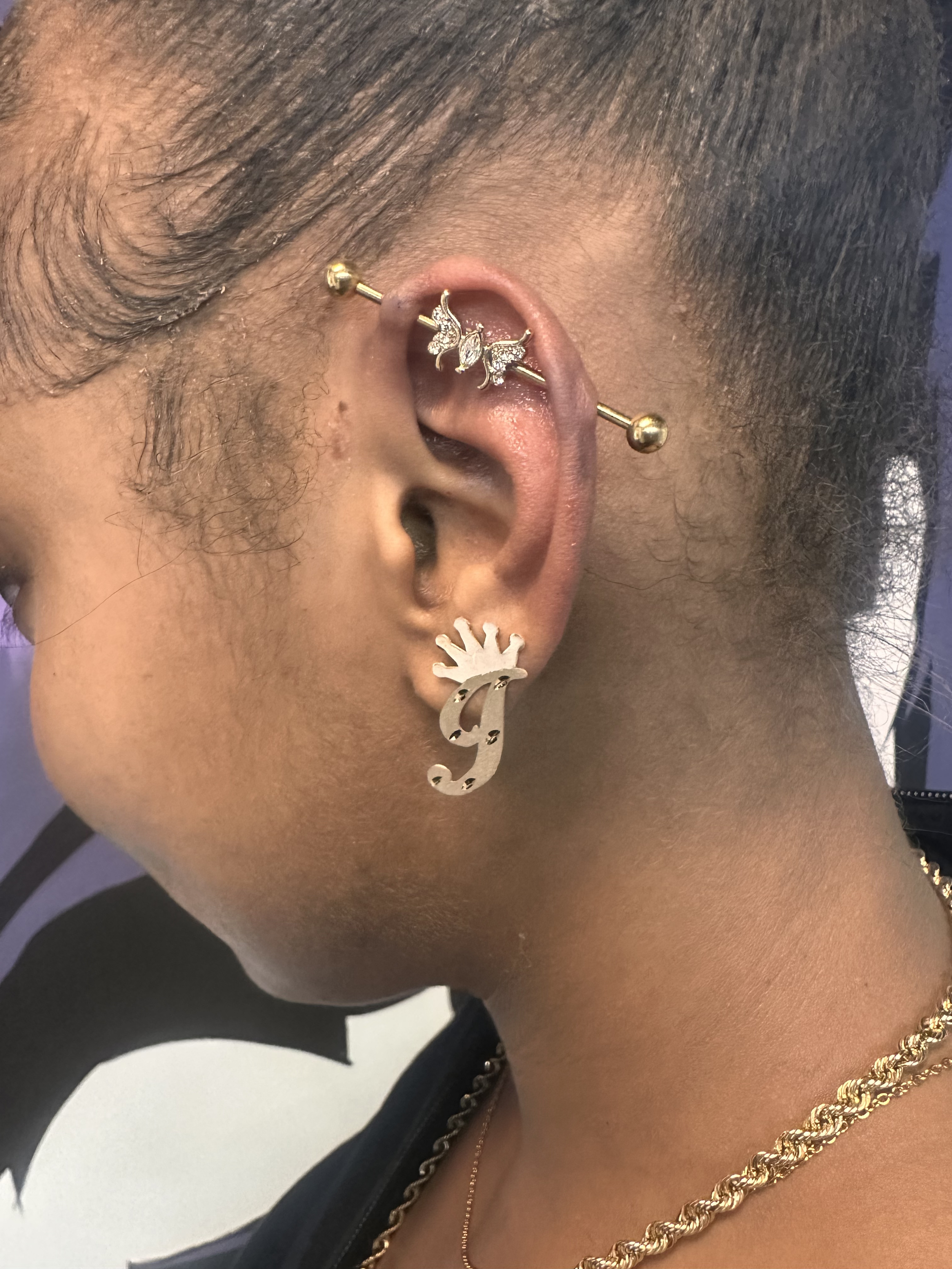 Close-up of a person's ear with multiple earrings, including a gold hoop with an intricate design, a gold barbell, and a large gold earring with a stylized letter 'Y' in it.