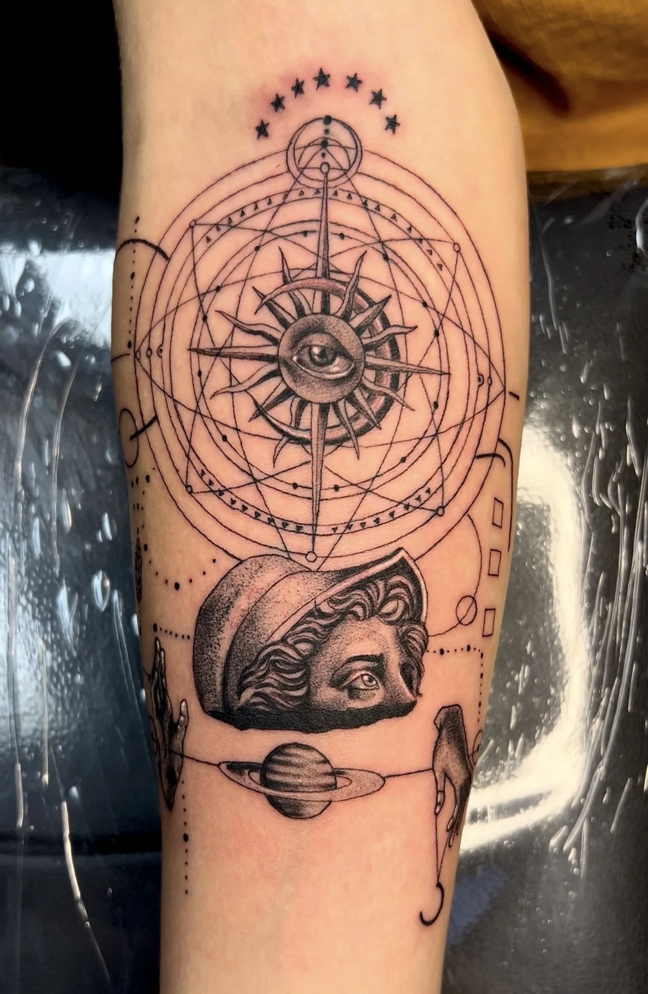 Tattoo featuring an eye in the center of a star with radiating shafts, surrounded by geometric lines and patterns, a woman’s face with a hat, a planet with rings, and a hand.