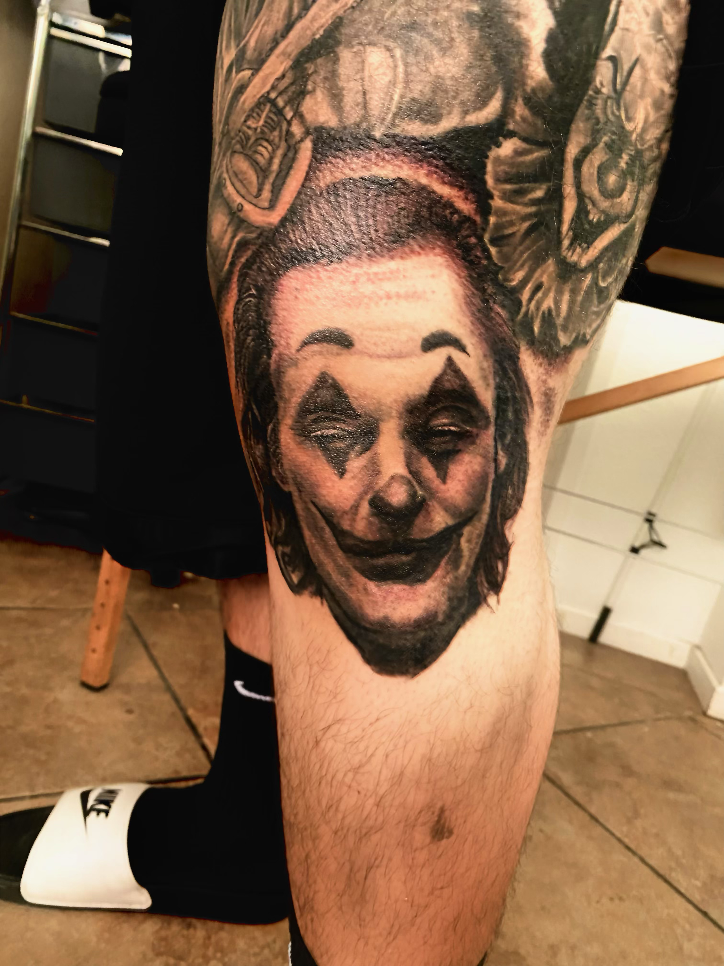 Tattoo of a smiling clown face on a person's thigh, with exaggerated makeup and dark eye makeup.