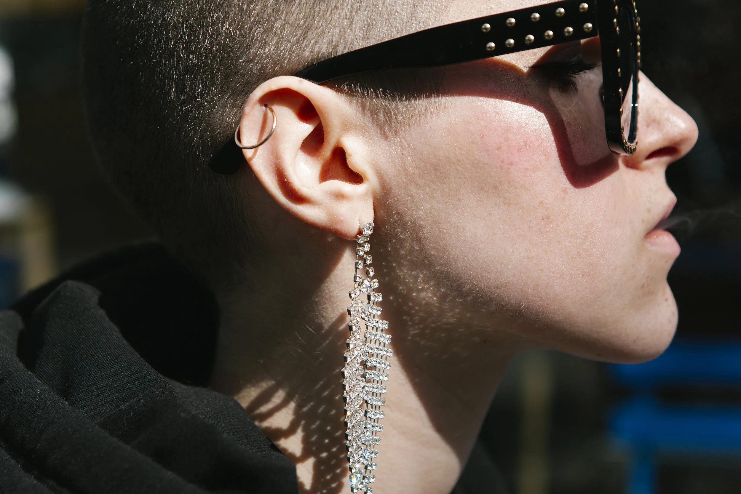 Close-up of a person's face with dark sunglasses, multiple earrings, and a large dangling diamond earring, showing side profile with short hair and part of their lips.