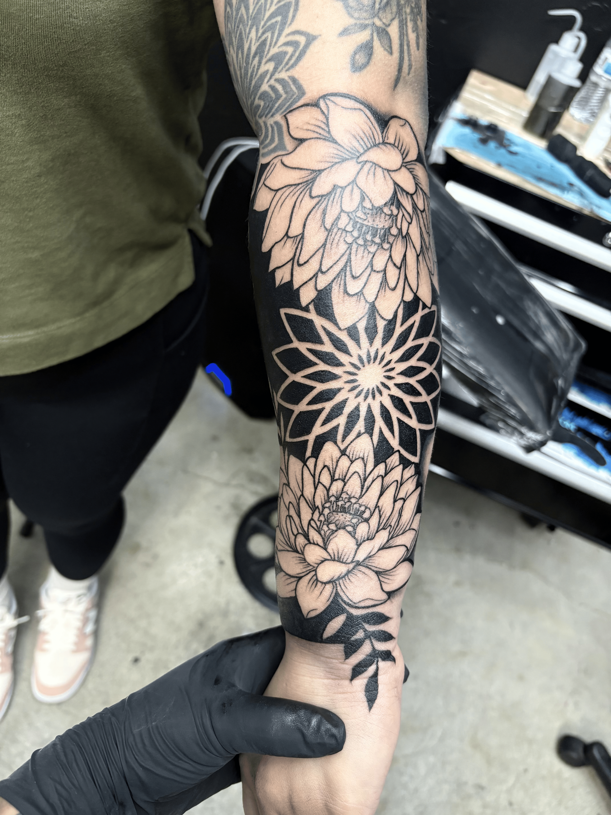 A person's arm with a tattoo of large flowers and intricate geometric patterns, being held by a tattoo artist wearing a black glove.