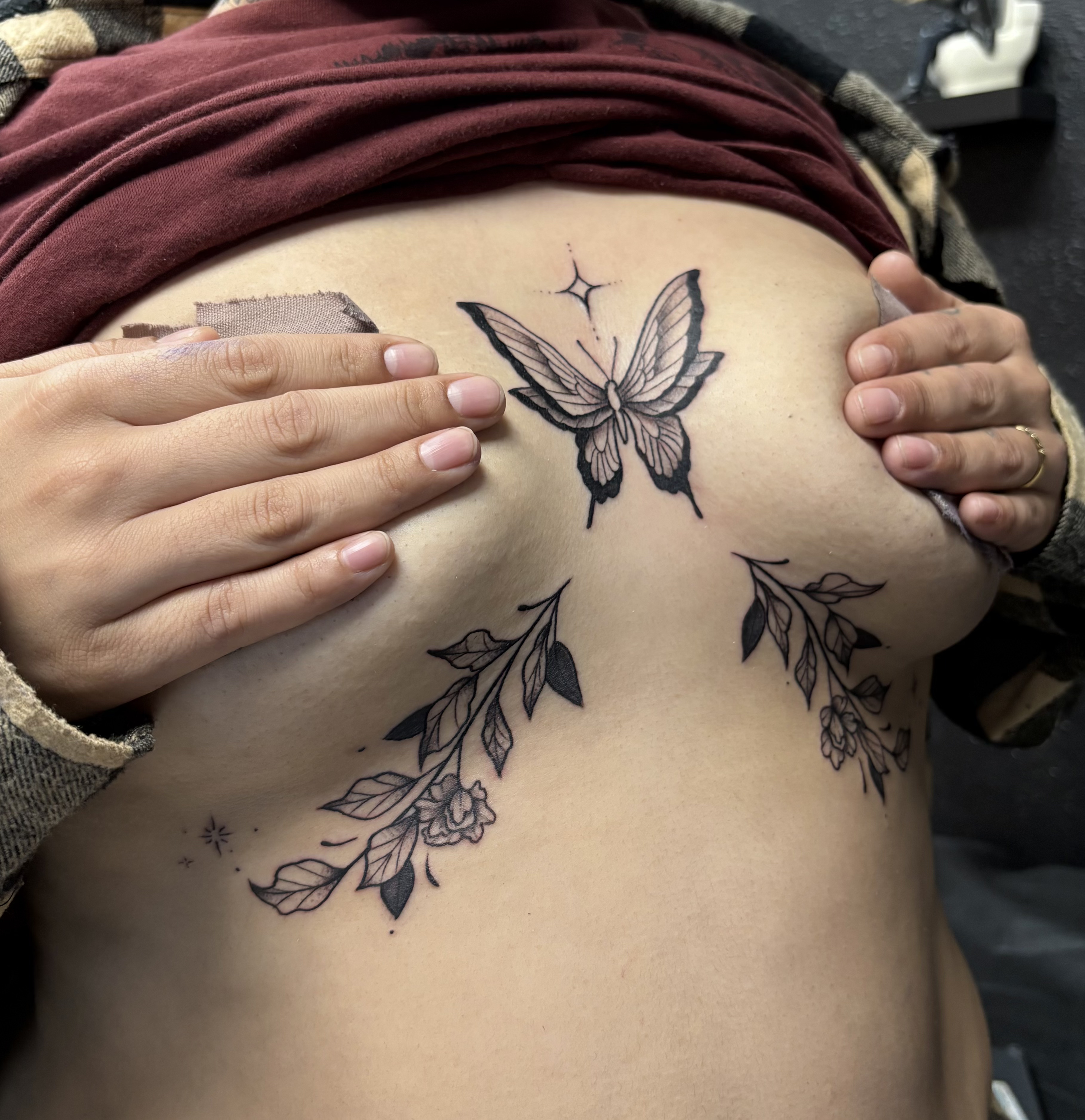 A person holding their shirt to reveal a tattoo of a butterfly with floral and leaf designs on their chest.