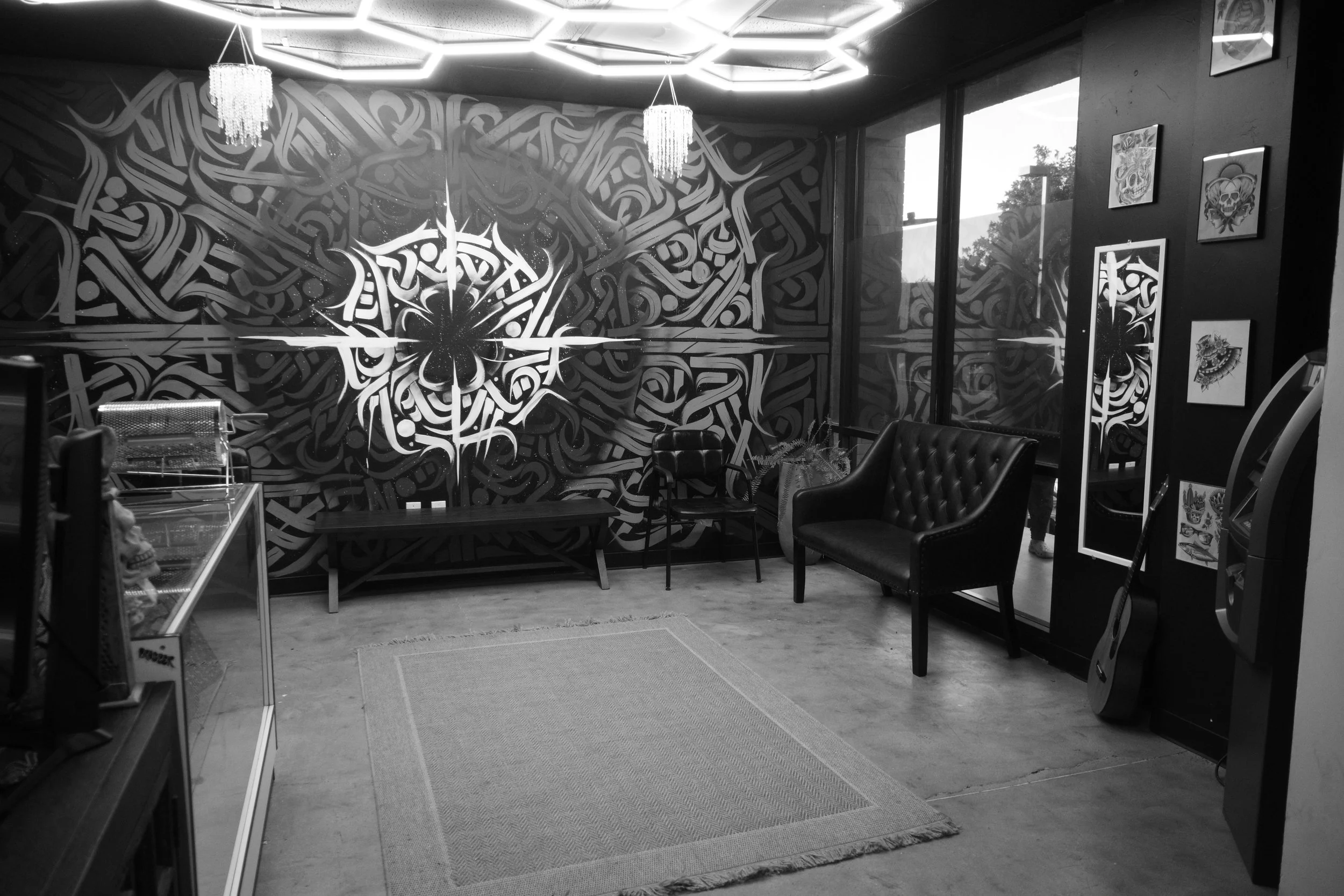 A waiting area with a textured, patterned wall, two chandeliers, a wooden bench, two chairs, a potted plant, a mirror, and artwork on the wall, with large windows showing trees outside.