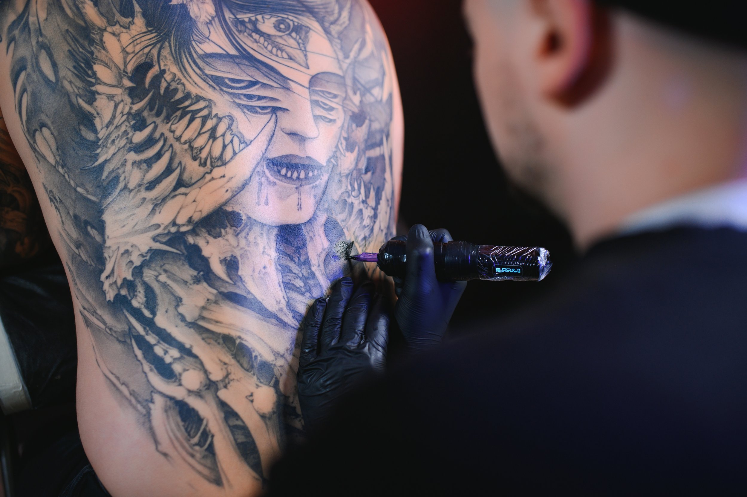 A person with a large, intricate tattoo on their upper arm is getting tattooed by an artist wearing black gloves. The tattoo features a detailed portrait of a creature with dragon-like elements and a human face with intense eyes and open mouth.