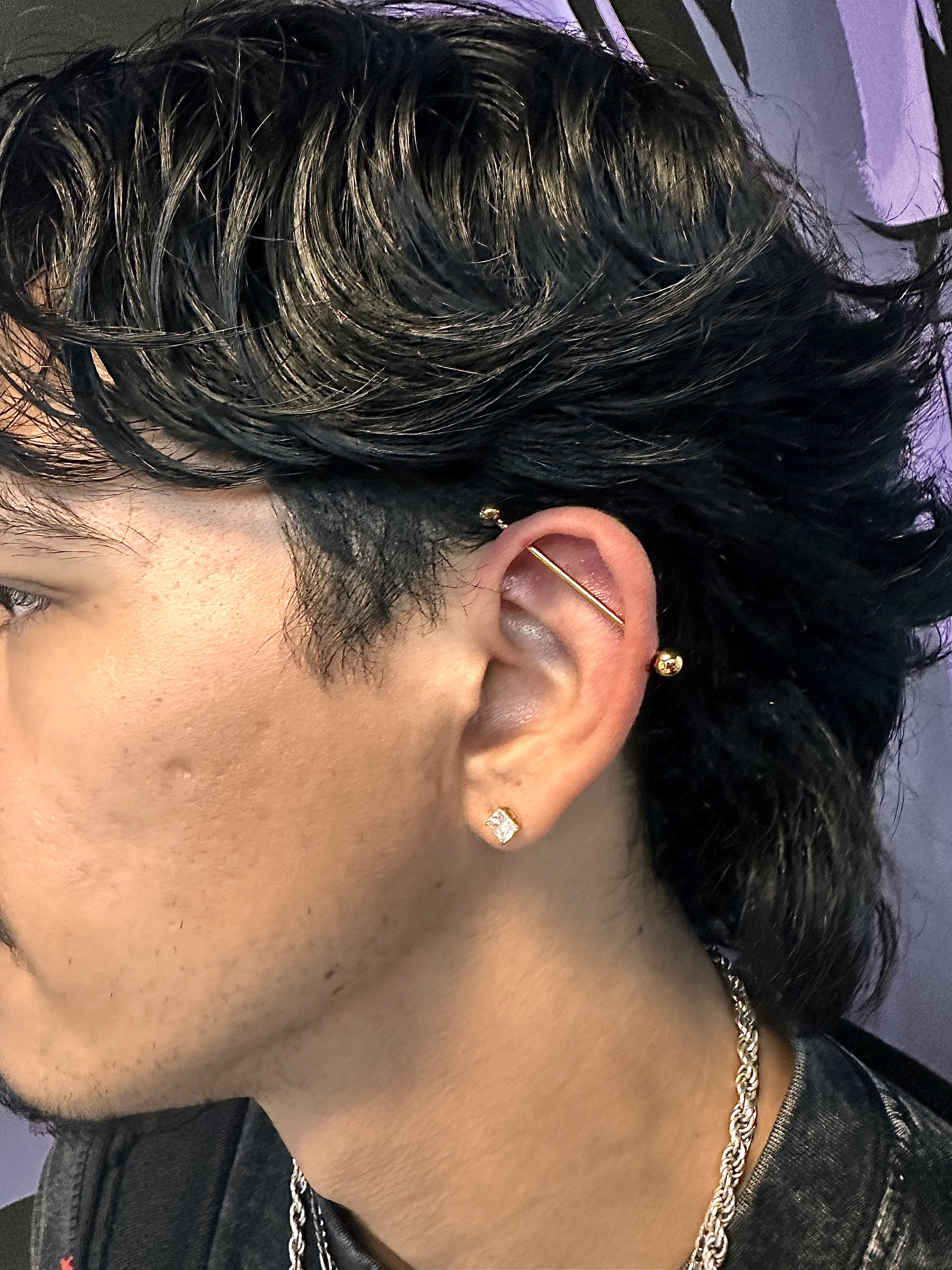 A young person with styled black hair, multiple earrings including a diamond stud, a gold bar hairpin, and a gold ball, wearing a chain necklace and black clothing.