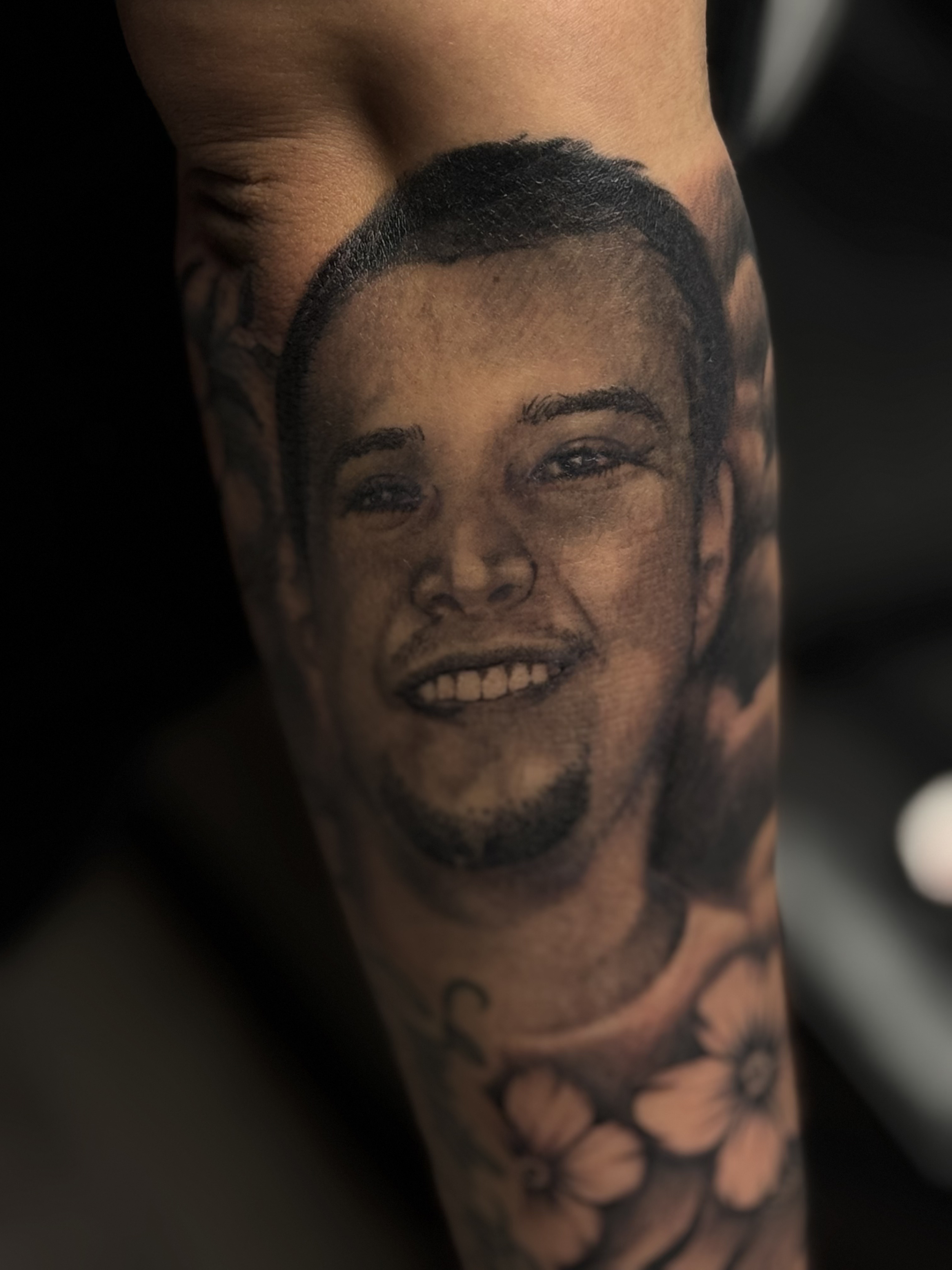 Tattoo of a smiling man's face with short hair on a person's arm, with flowers below the face.
