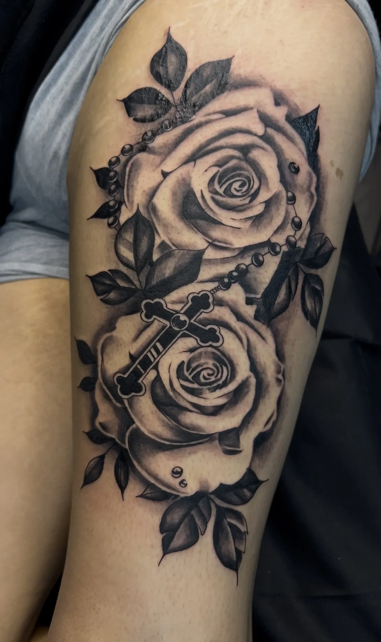 Black and gray tattoo of two roses, leaves, a rosary, and a cross on a person's upper arm.