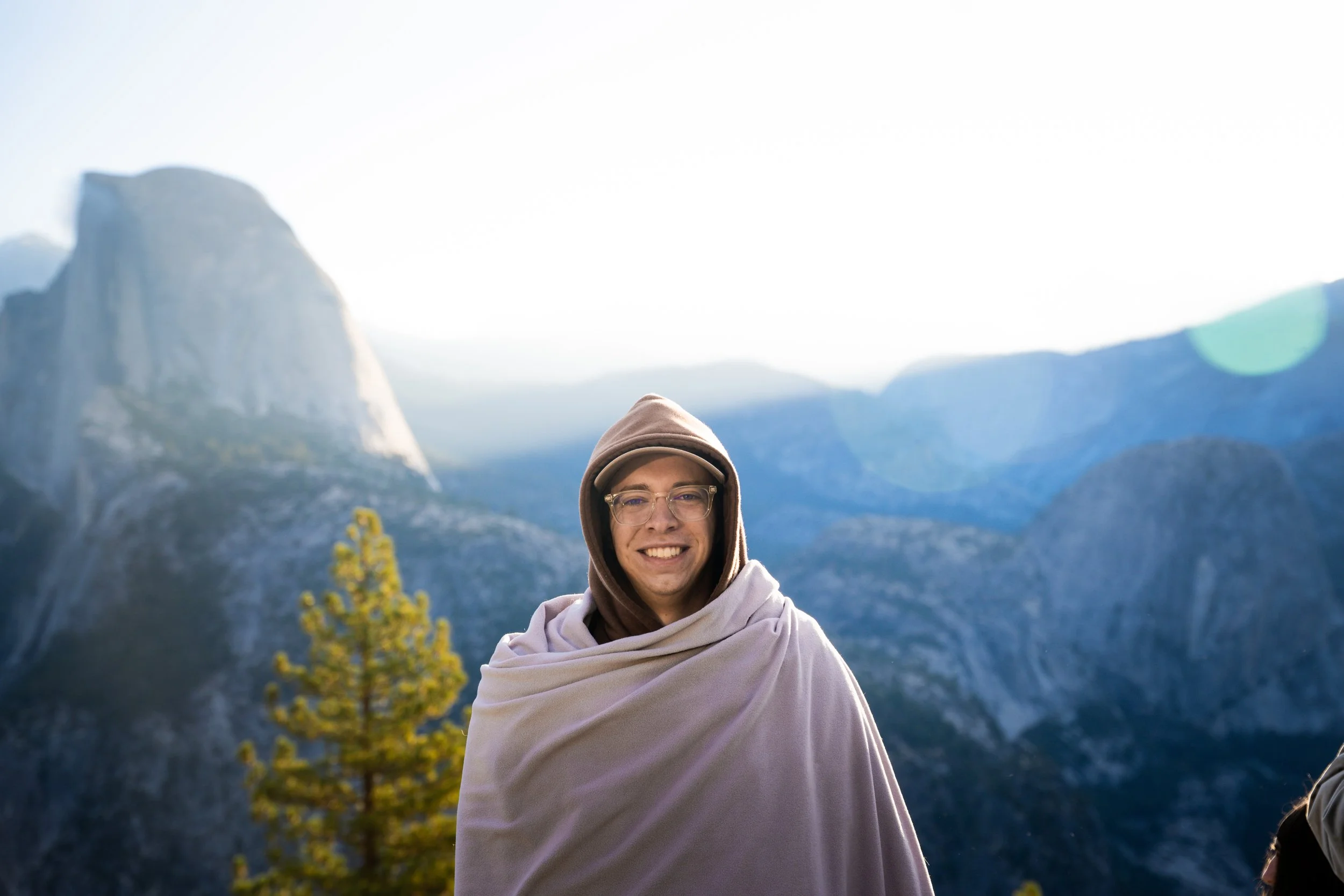 A smiling woman wearing glasses, a hoodie, and a blanket standing outdoors in a mountainous area with large rock formations and trees in the background.