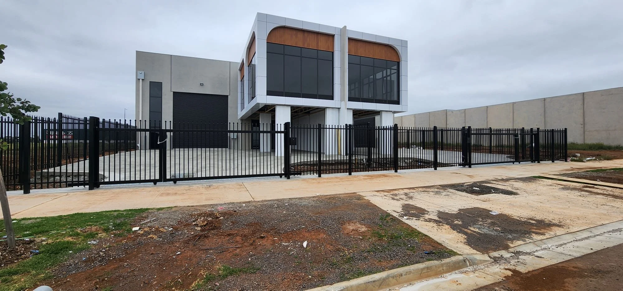 Modern two-story house with large glass windows, white walls, and wooden accents, surrounded by a black metal fence, with a concrete driveway and some patches of grass in the foreground