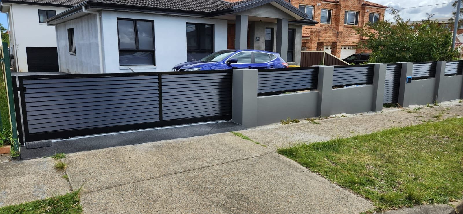 Modern house with a blue car parked in front, secured by a black metal and concrete fence with horizontal slats, sidewalk, and a grassy area.