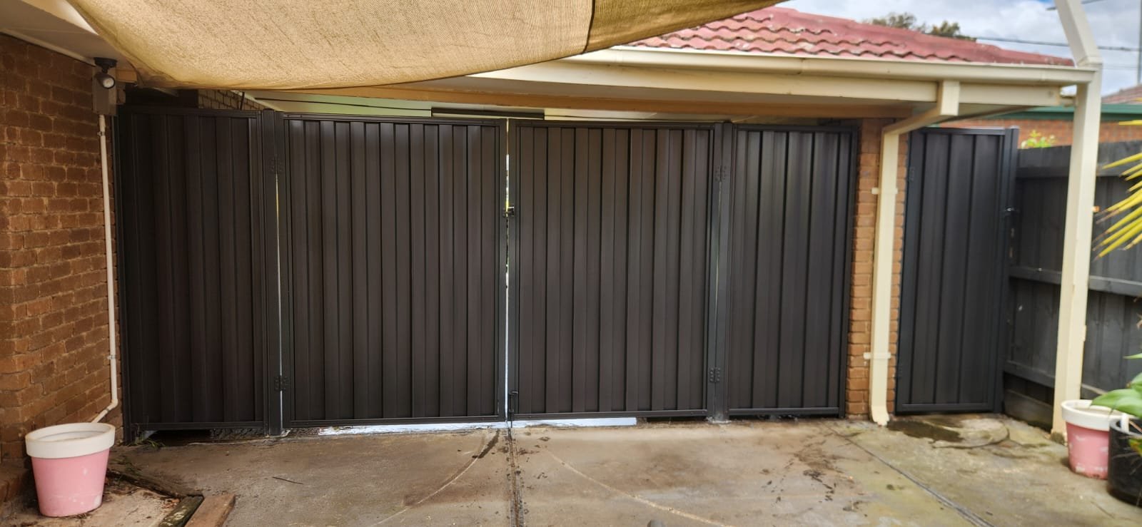 Black metal gate with double doors and a side panel on the right, set between brick walls, with concrete ground and some plant pots nearby.