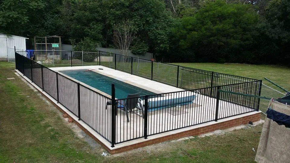 A backyard with a rectangular in-ground swimming pool, surrounded by a black metal fence, grass, and trees in the background.