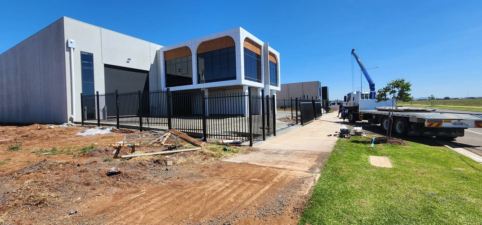 Under construction modern building with black fence, sidewalk, grass, construction truck, and crane under a blue sky.