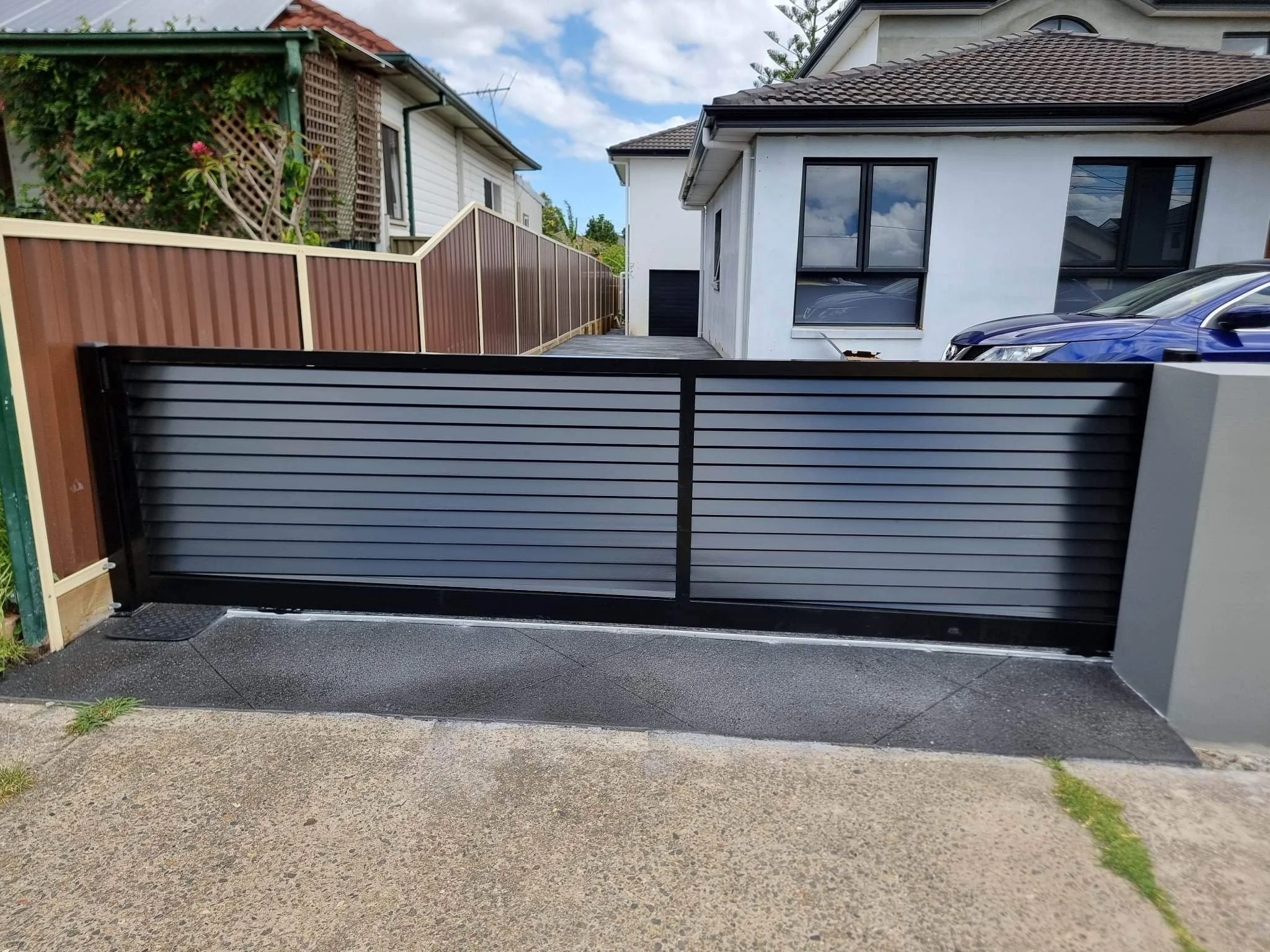 A modern black metal sliding gate in front of a driveway, with a concrete sidewalk in the foreground, a white house with dark window frames and a blue car parked beside it.