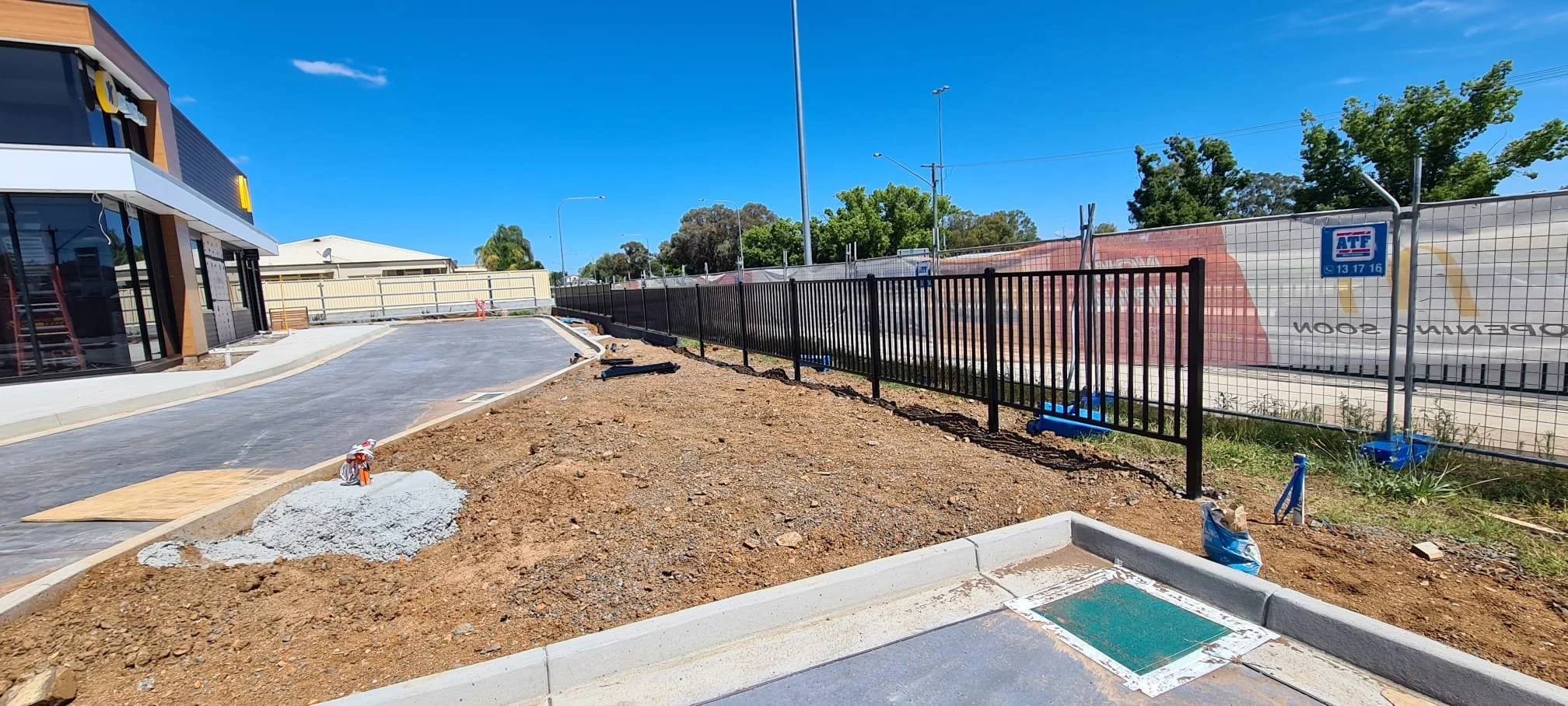 Construction site with fresh pavement, a black metal fence, and an outdoor area near a commercial building under a clear blue sky.