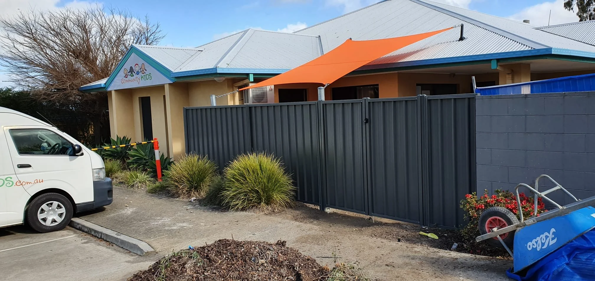 Exterior view of a community kids' center with a gray metal fence, white van, plants, and a building with a sign that says 'Community Kids'.