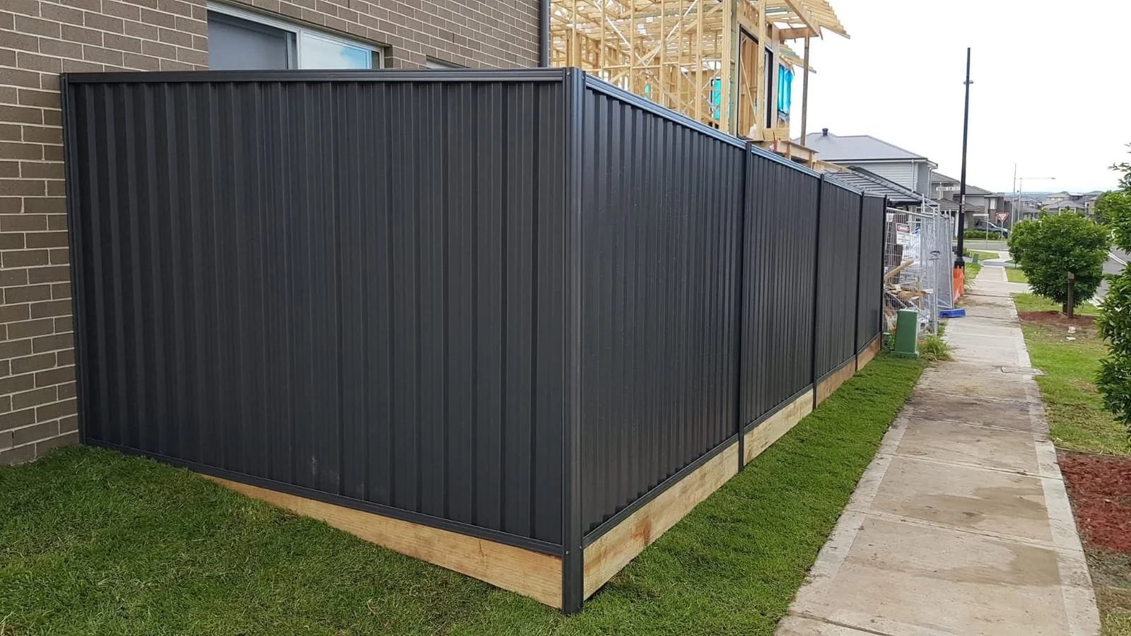 Black metal fence along sidewalk with grass and trees nearby, under construction house in background.