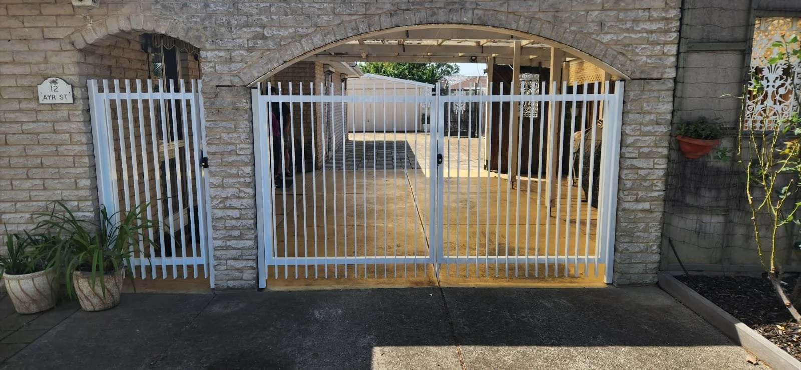 A white metal sliding gate in front of a brick house with an arched brick opening and a small side gate. Potted plants are on either side of the gates. The house number 12 and street name 'Ayr St' are visible on a sign to the left.
