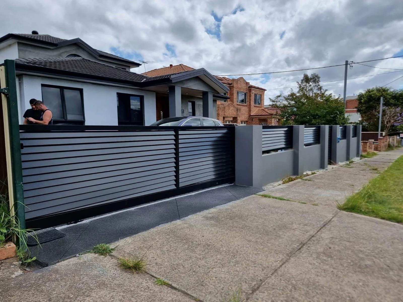 Photo of a modern house with a gray and black horizontal slat fence and a sliding gate, with a woman standing near the fence on the left side of the image.