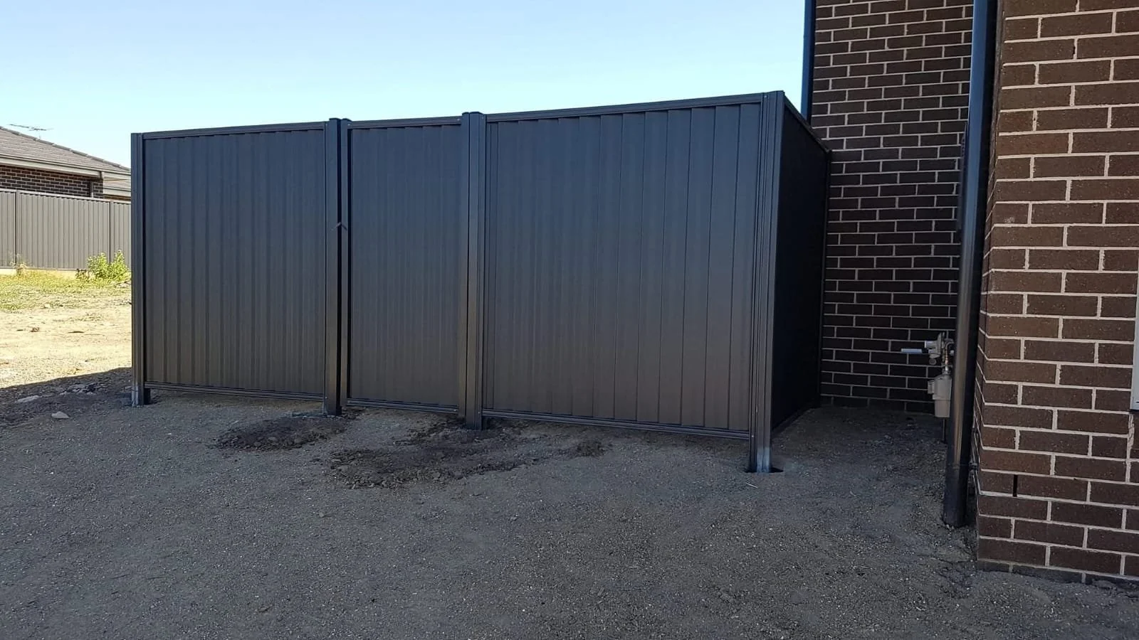 Gray metal fence panels with gate next to a brick wall with faucet and pipe, on a dirt ground outside under clear sky.