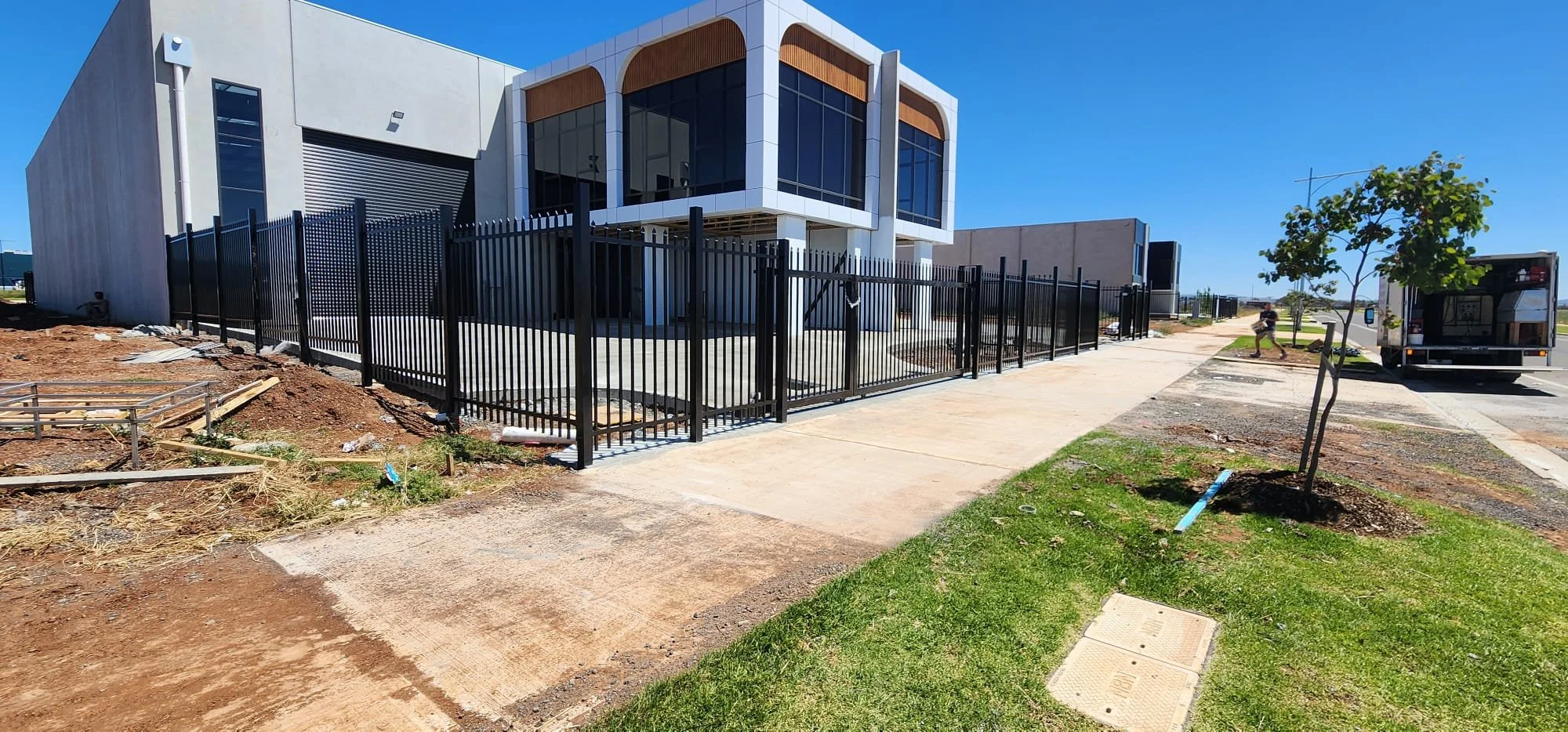 Modern building with large windows, surrounded by a black metal fence. Construction work visible with dirt and building materials on the ground. Sidewalk and green grass with small trees along the street, blue sky.