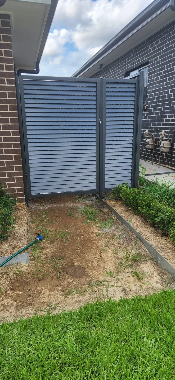 Newly installed gray louvered privacy screen on a small lawn area next to a brick house with some garden plants and a garden hose on the ground.