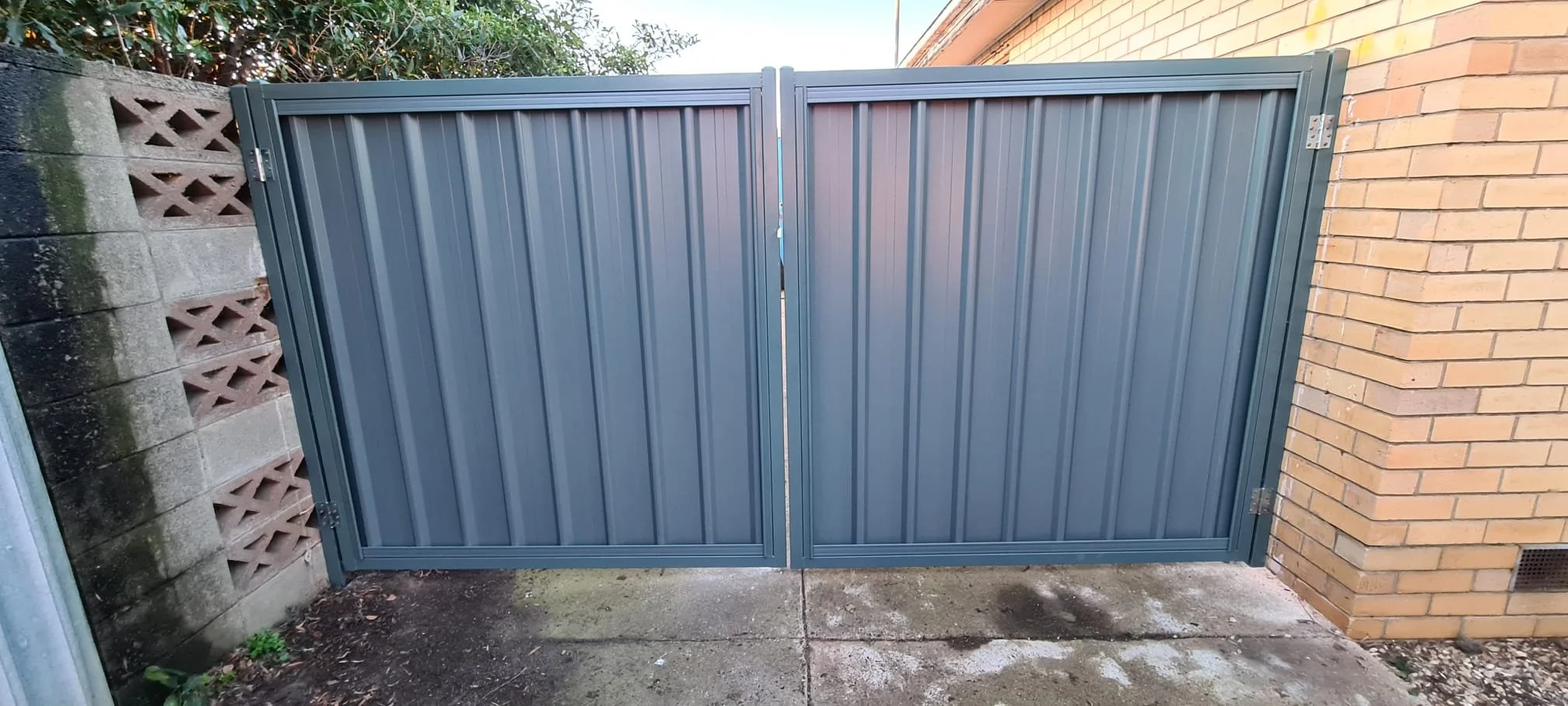 A blue metal gate with vertical ridges, closed, situated between a concrete block wall and a brick wall, on a concrete ground.